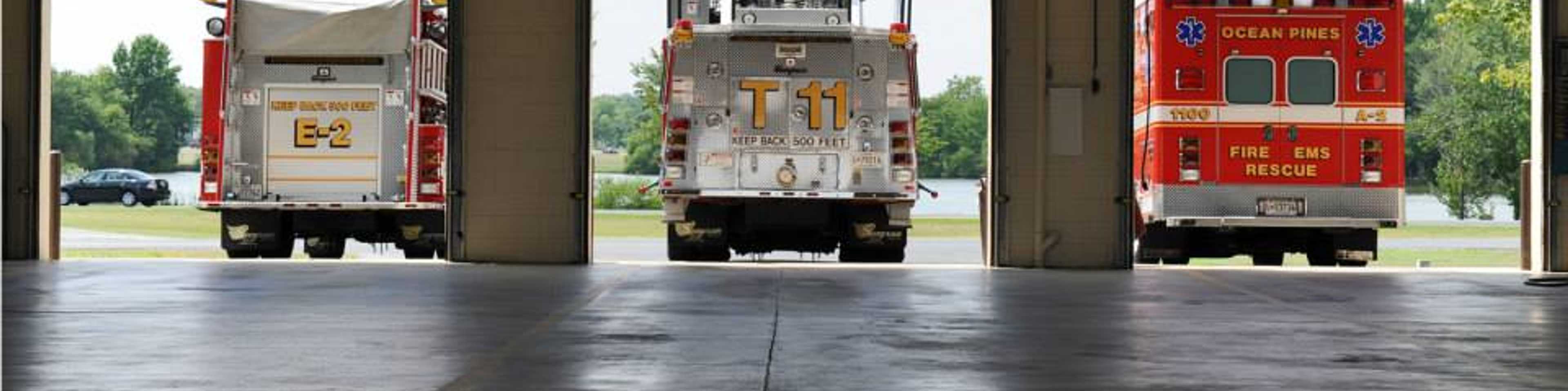 Three fire trucks parked in open bay doors of a fire station, with trees and sky visible outside.