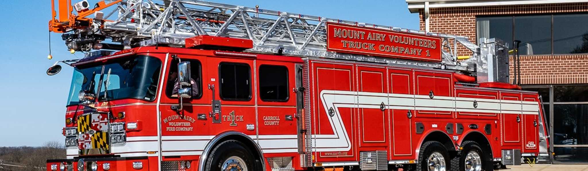 A red and white Mount Airy Volunteers fire truck with an extended ladder is parked on a concrete driveway next to a brick fire station building under a clear blue sky.