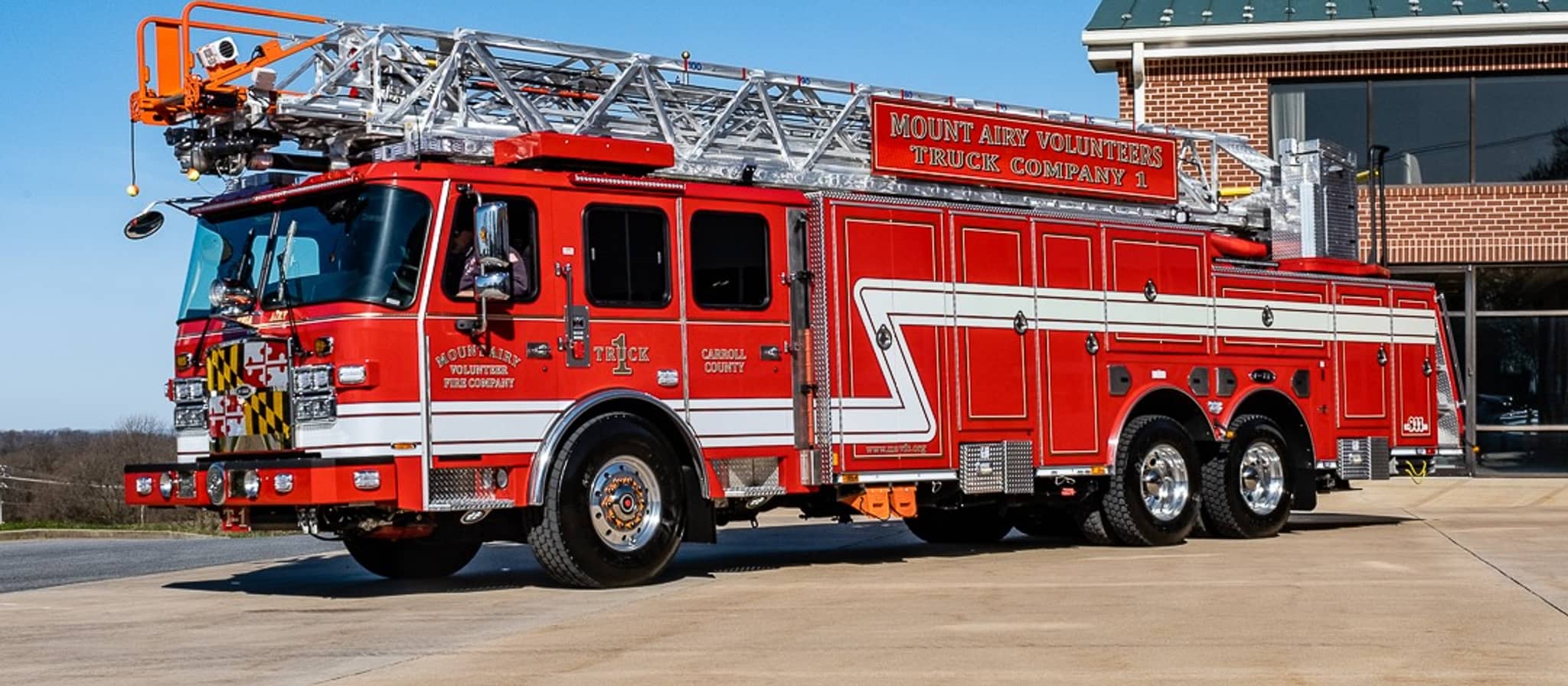 A red and white Mount Airy Volunteers fire truck with an extended ladder is parked on a concrete driveway next to a brick fire station building under a clear blue sky.