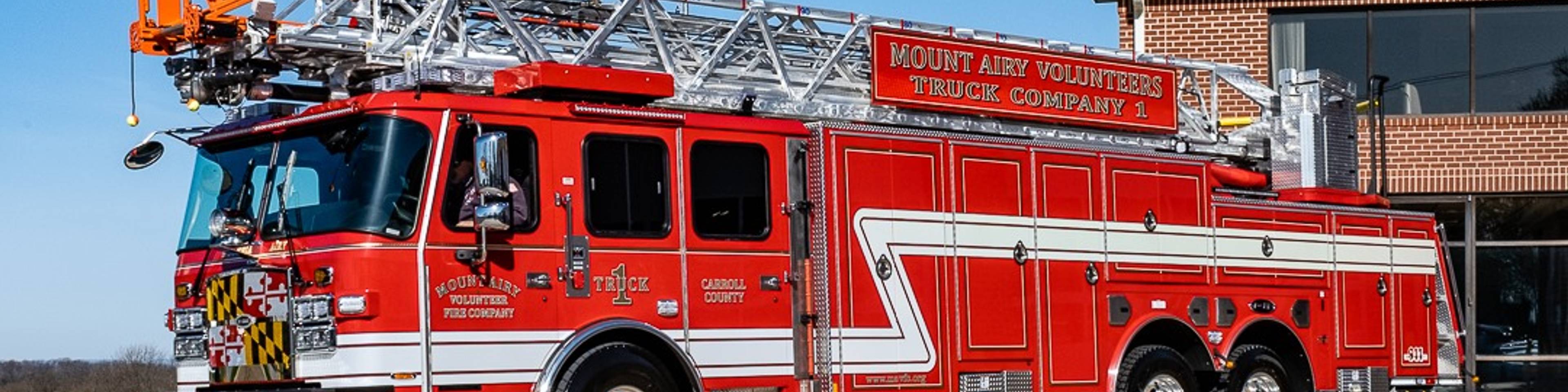 A red and white Mount Airy Volunteers fire truck with an extended ladder is parked on a concrete driveway next to a brick fire station building under a clear blue sky.