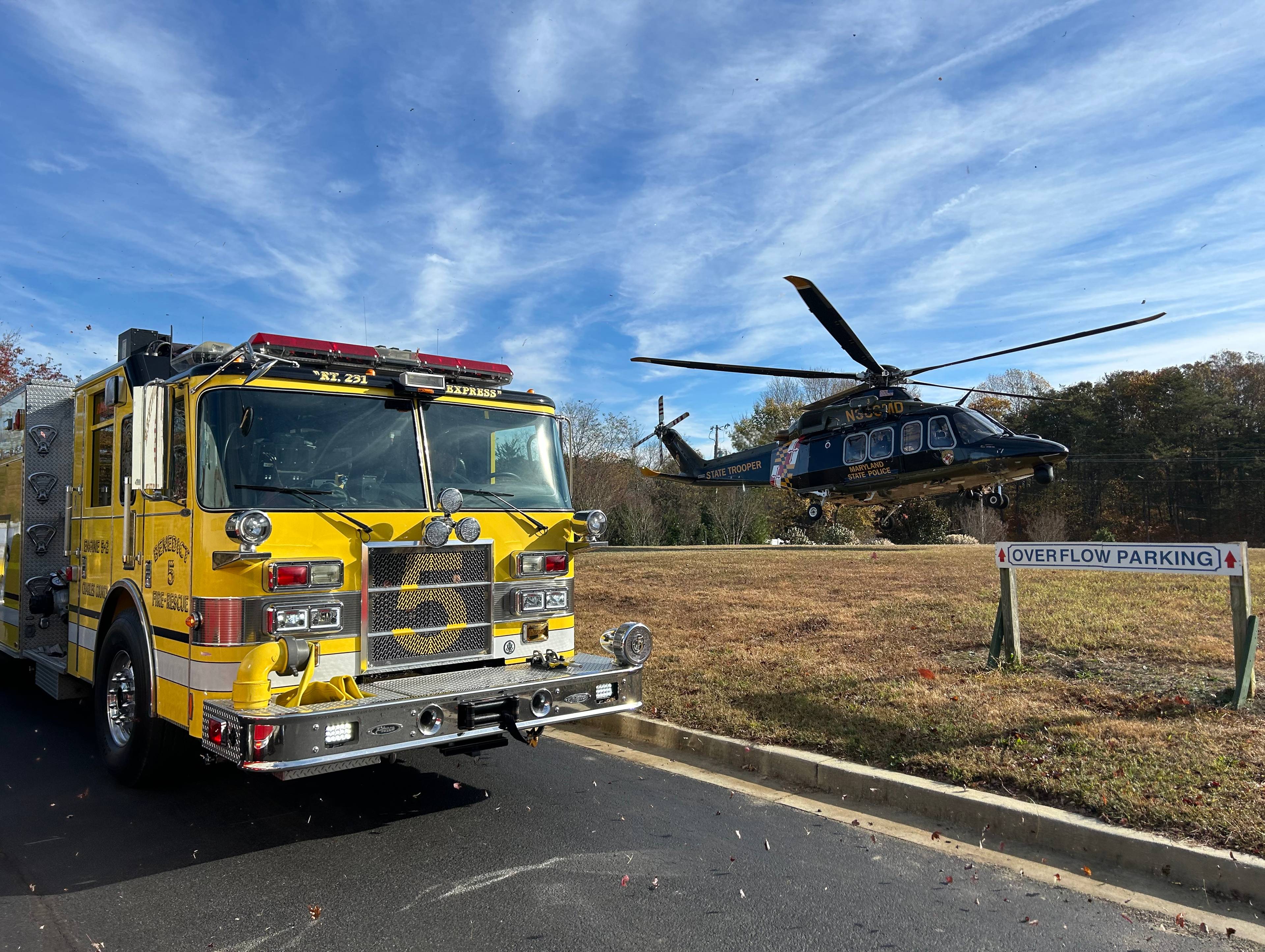 A yellow fire truck parked on a road beside a dry grass field with a state trooper helicopter landing in the background under a blue sky with wispy clouds.