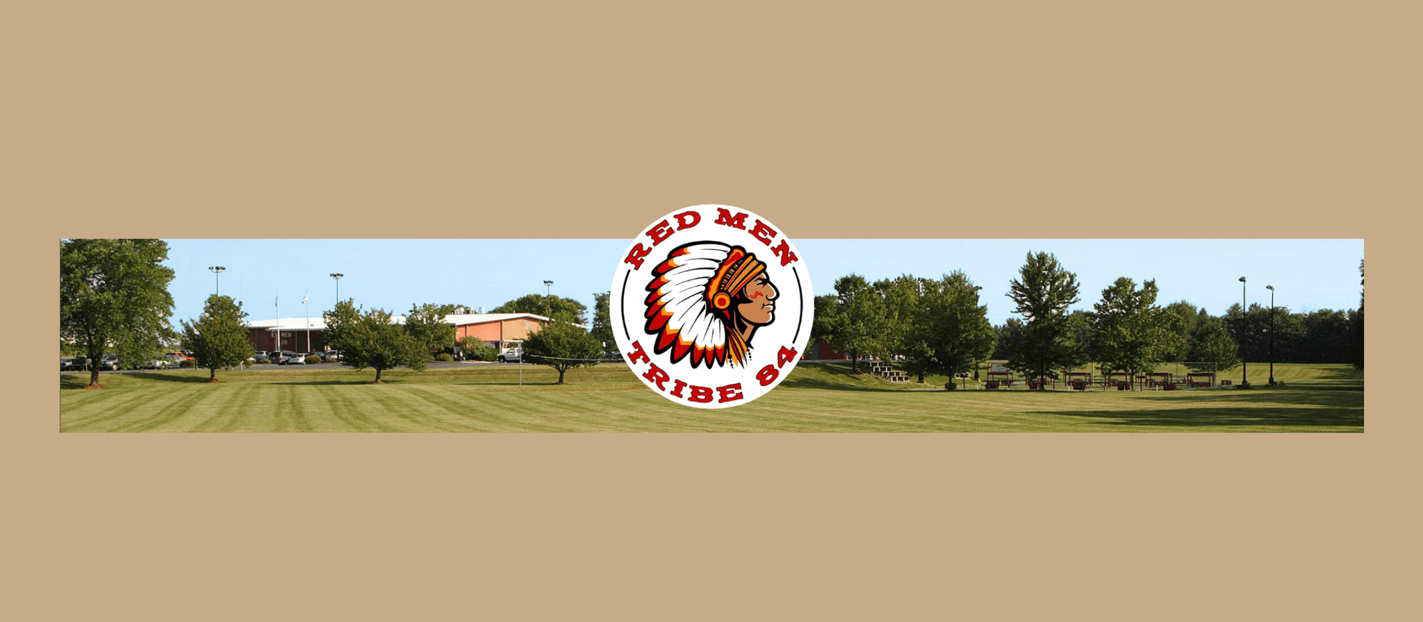 A wide panoramic view of a sports field with manicured grass, trees, and school buildings in the background. The Red Men Tribe 84 logo featuring a Native American chief's head in a circle is overlaid in the center.