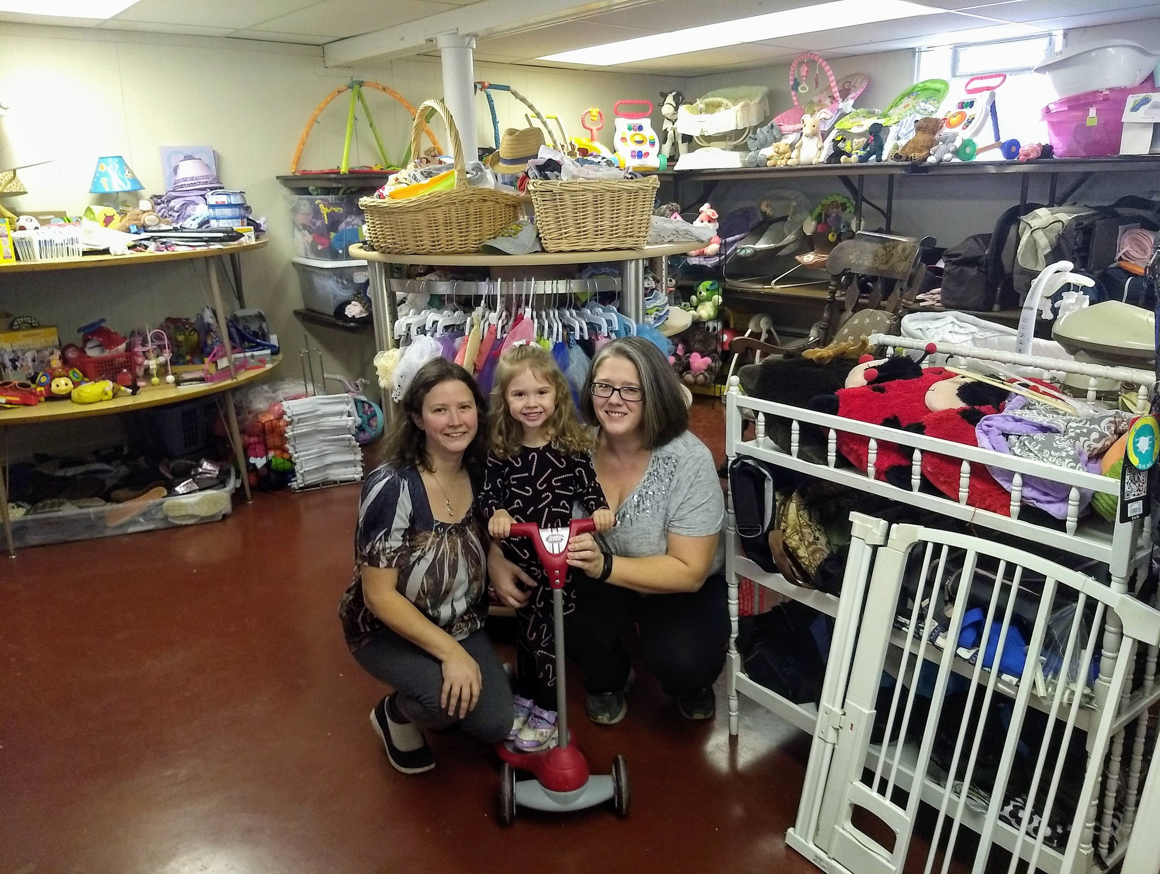 Three smiling women and a young child posing together in a cluttered storage room filled with toys, clothing, baskets, and household items on shelves and racks.
