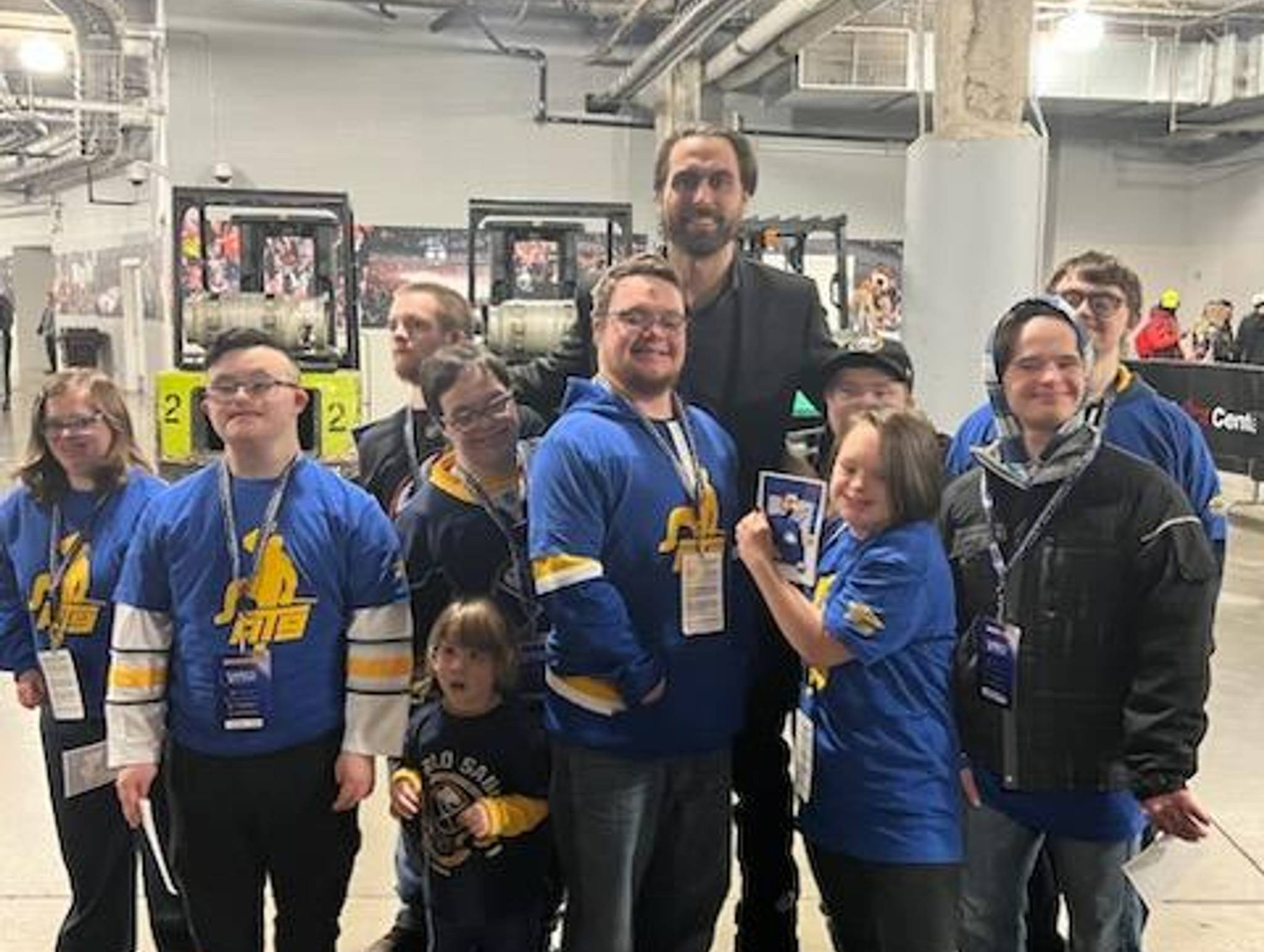 A group of people, including children and adults wearing blue and yellow team apparel with lanyards, pose together in an industrial warehouse or event space with exposed ceiling infrastructure and overhead lighting.