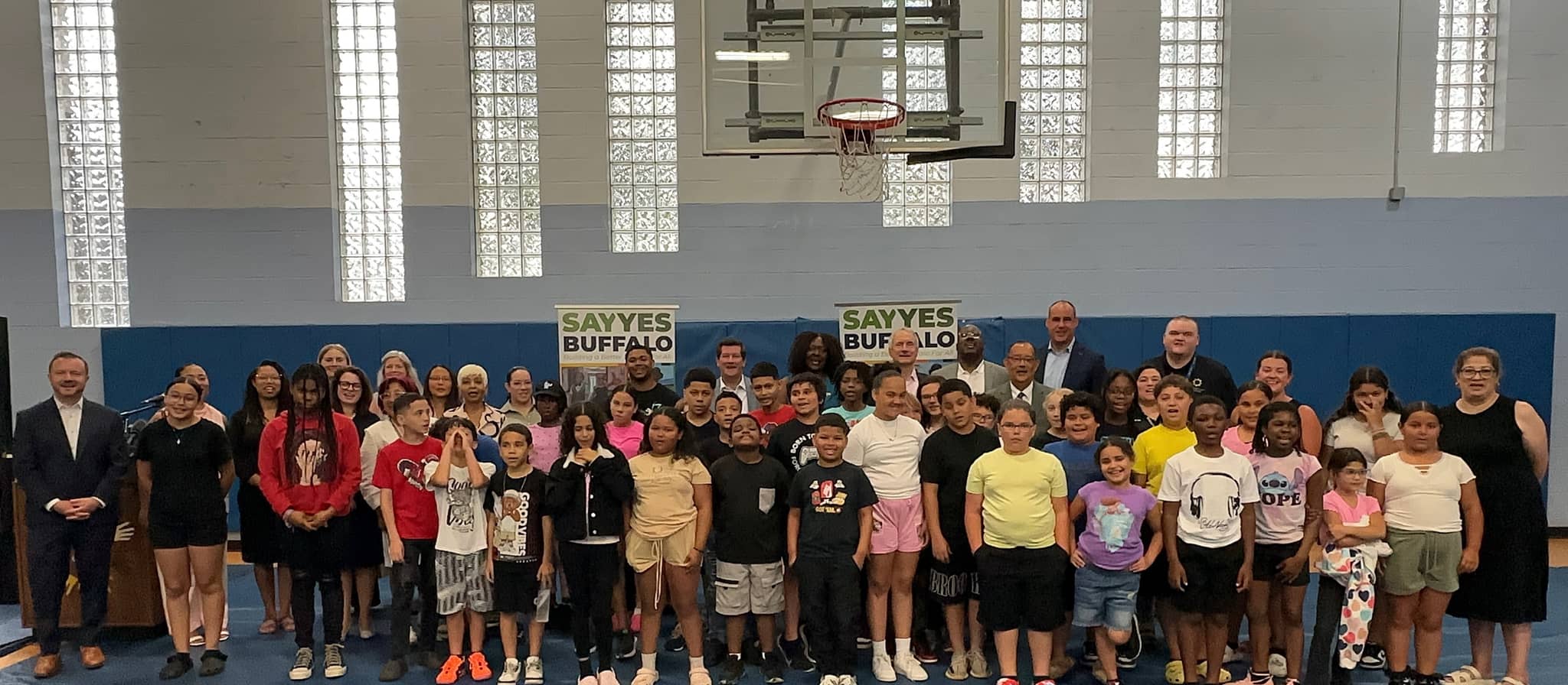 Large group of children and adults standing together on a blue gymnasium floor in front of a blue wall with "Sayyes Buffalo" signs, with a basketball hoop visible above.