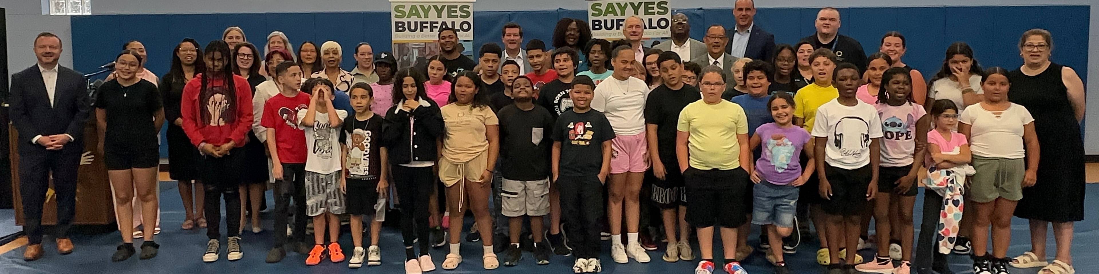 Large group of children and adults standing together on a blue gymnasium floor in front of a blue wall with "Sayyes Buffalo" signs, with a basketball hoop visible above.