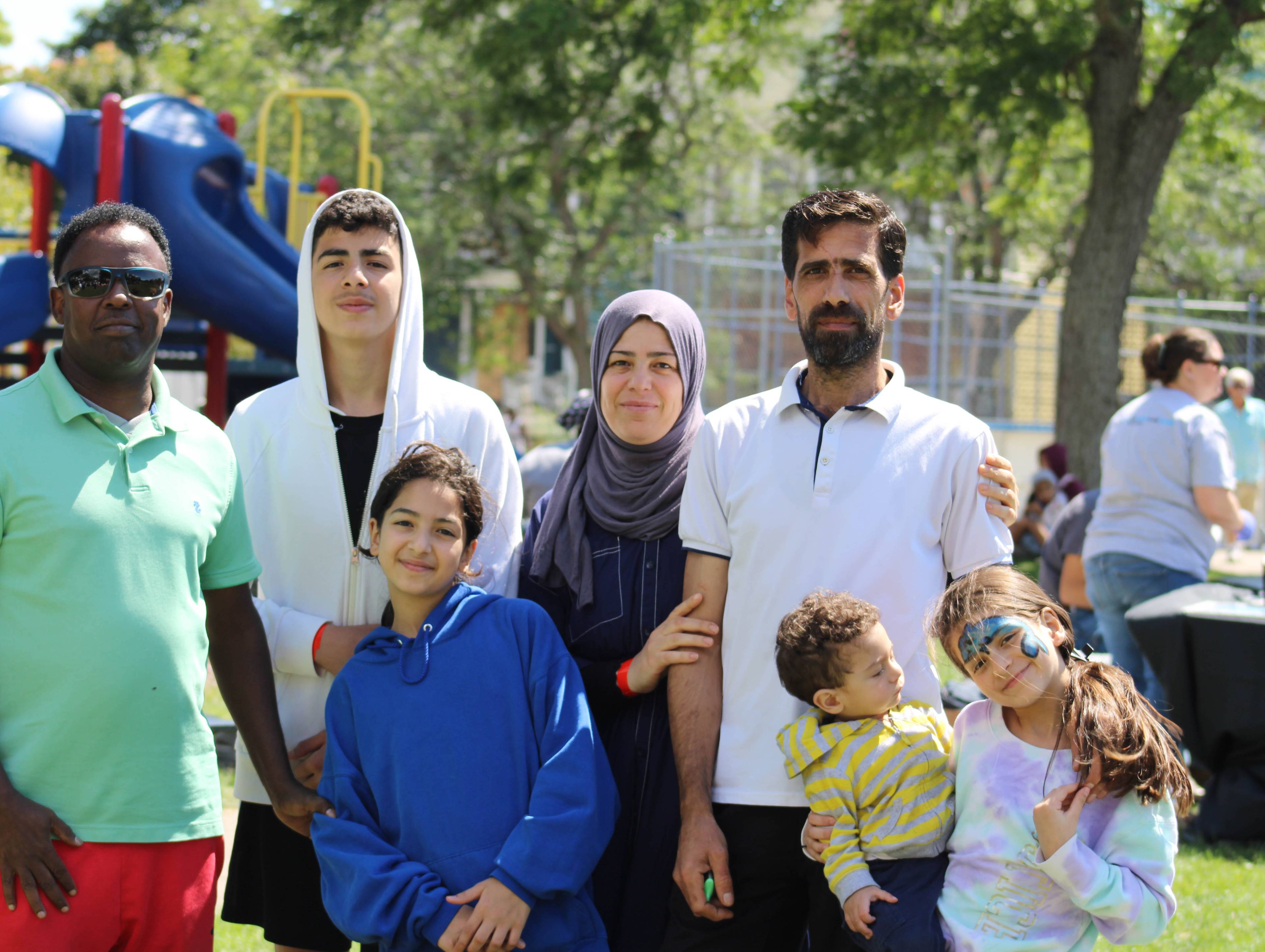 A diverse multi-generational family group stands together at a sunny playground, with children and adults of various ages smiling at the camera, trees and play equipment visible in the background.