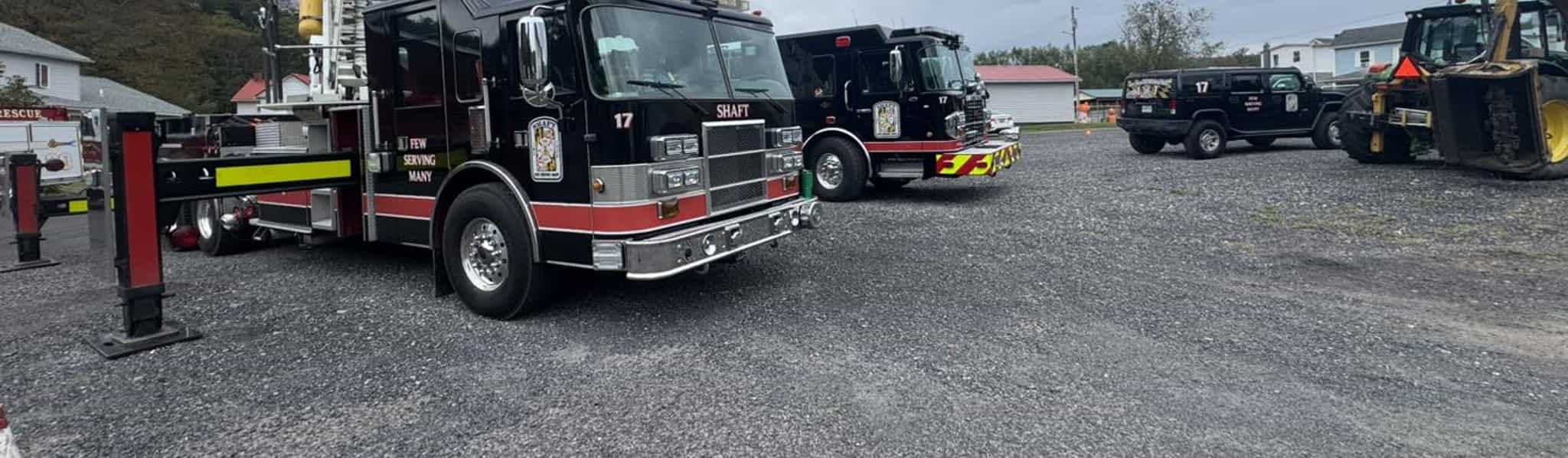 Fire department vehicles parked at a station lot, including a ladder truck with extended aerial ladder and other emergency response trucks under cloudy sky.