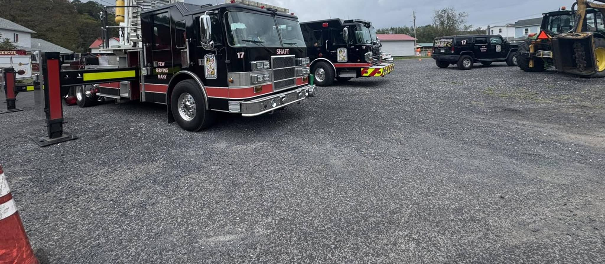 Fire department vehicles parked at a station lot, including a ladder truck with extended aerial ladder and other emergency response trucks under cloudy sky.