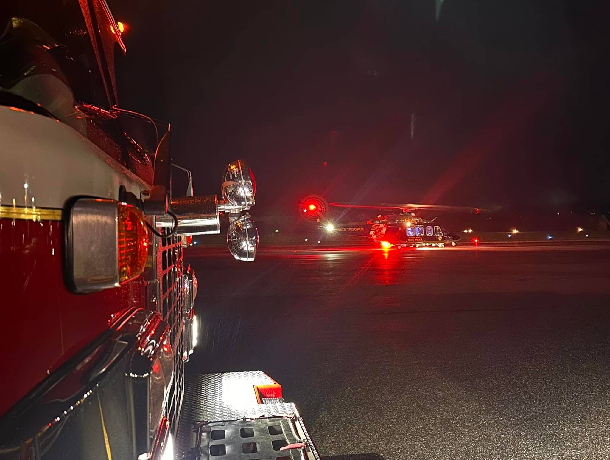 Fire truck at night with illuminated red and white lights parked on wet pavement, showing the truck's cab and equipment, with another emergency vehicle visible in the distance.