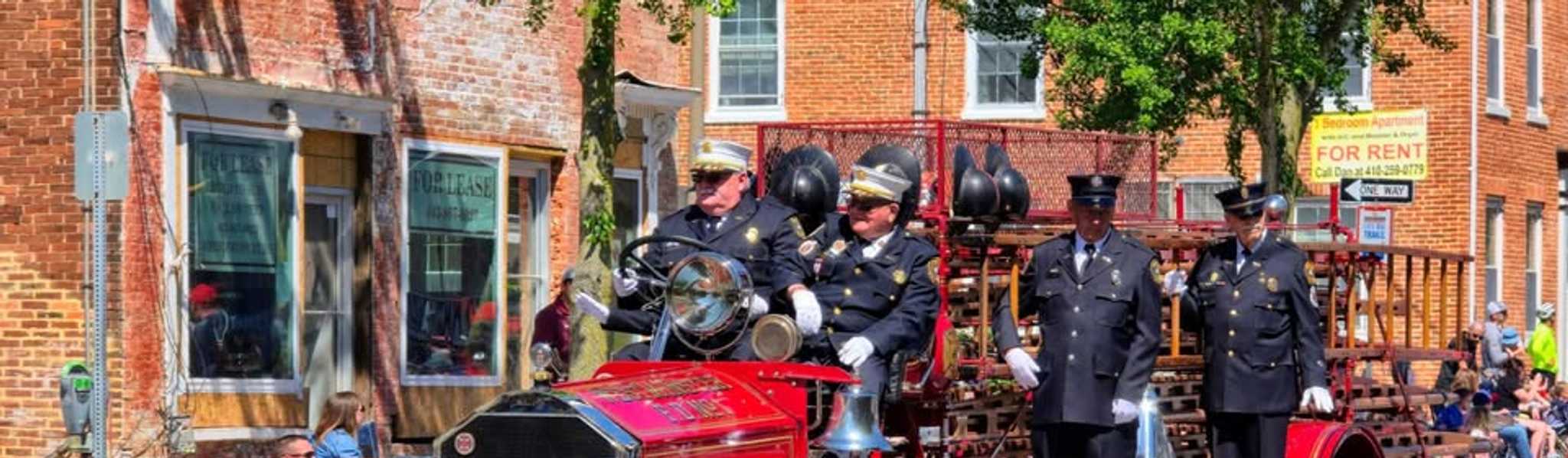 Vintage red fire truck with uniformed firefighters parading down a brick street lined with brick buildings and spectators sitting on the sidewalk.
