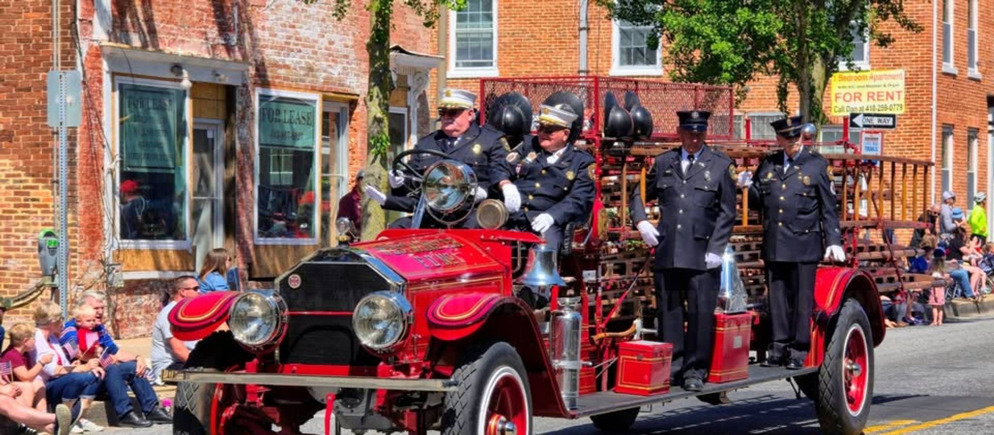 Vintage red fire truck with uniformed firefighters parading down a brick street lined with brick buildings and spectators sitting on the sidewalk.