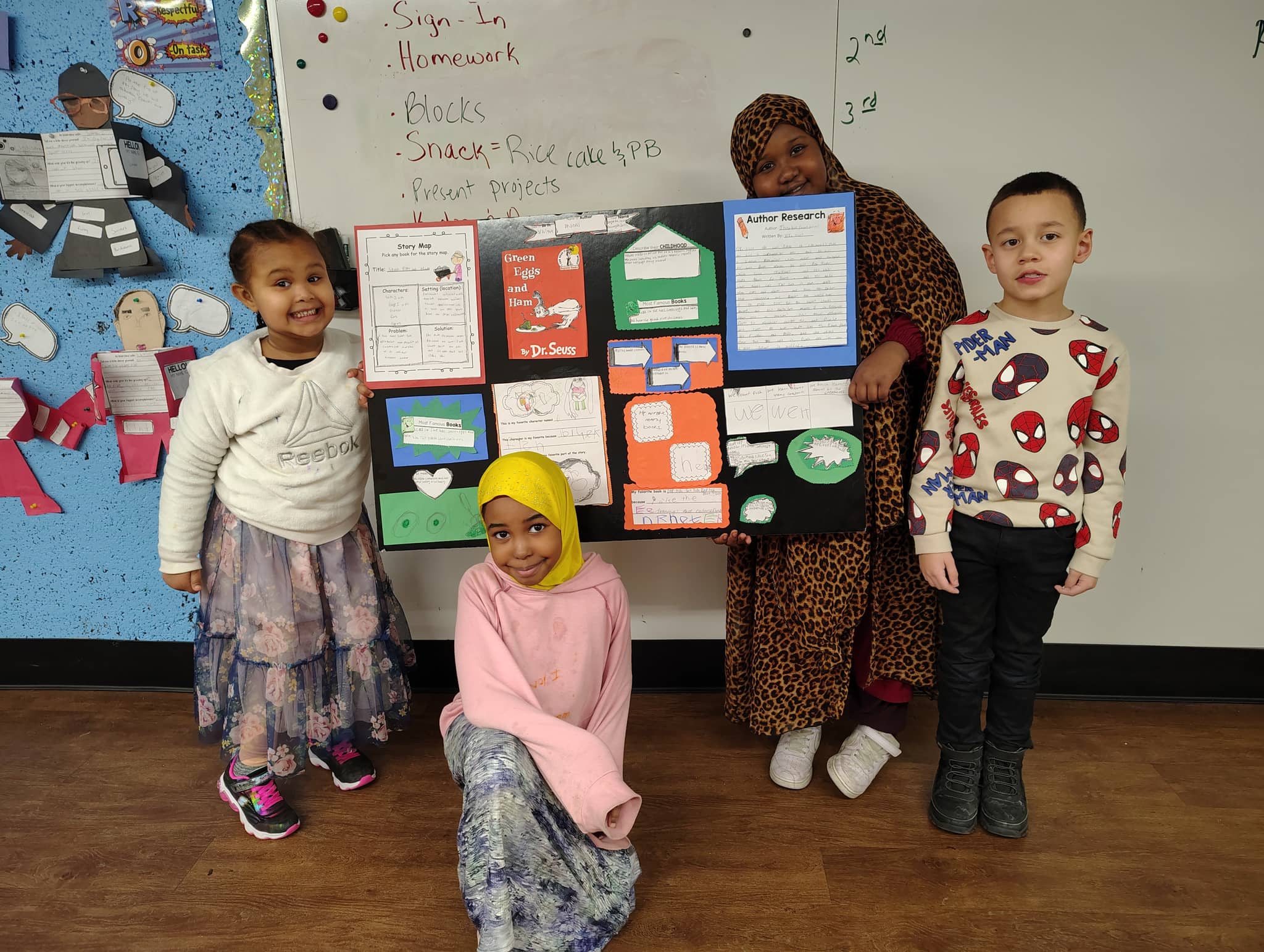 A teacher and four young students pose together in a classroom in front of a bulletin board displaying colorful student projects and worksheets related to books and learning activities.