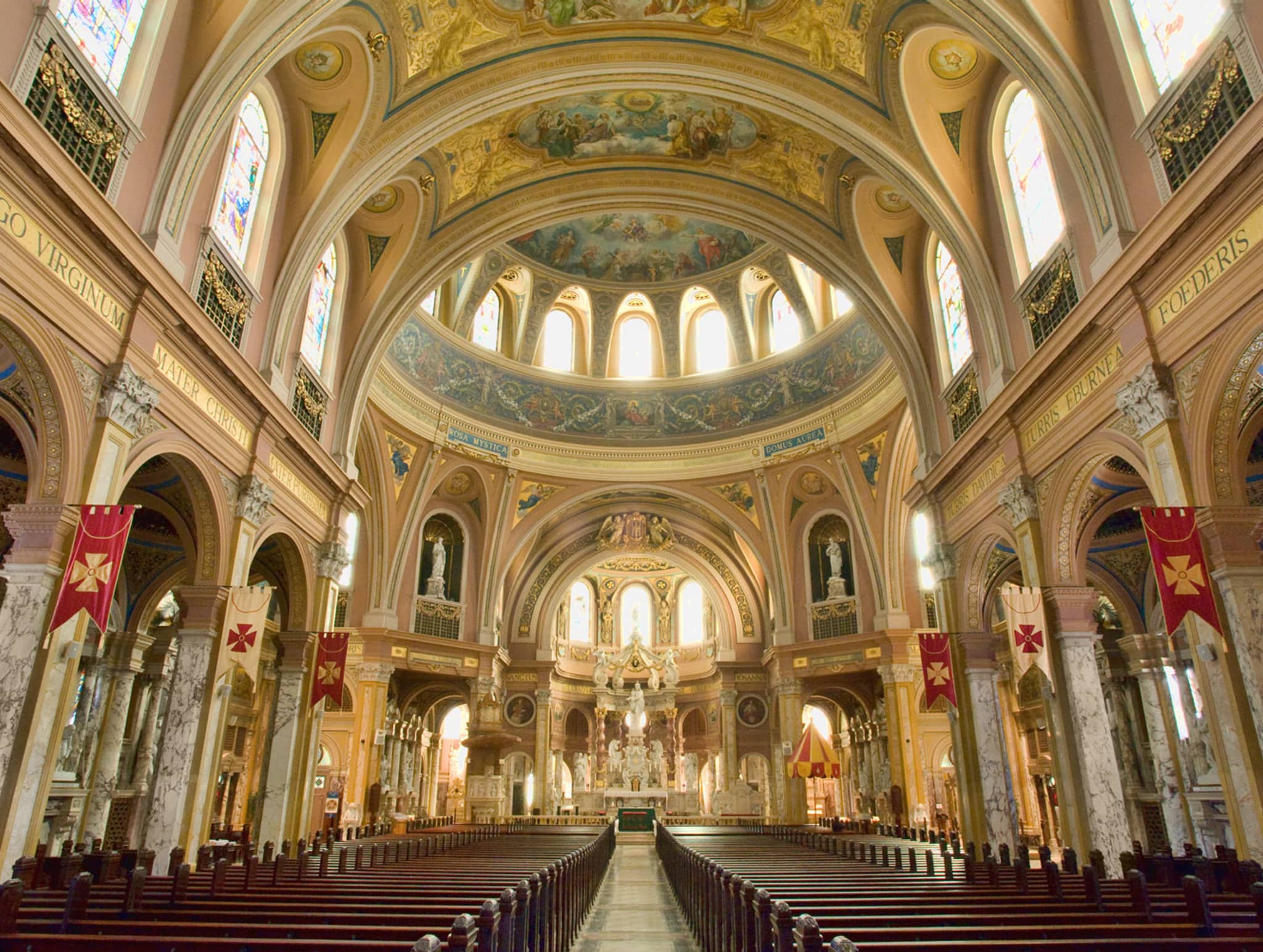 Interior of a grand ornate cathedral with a soaring domed ceiling, decorated arches, columns, stained glass windows, red banners, and wooden pews lining the nave toward the altar.