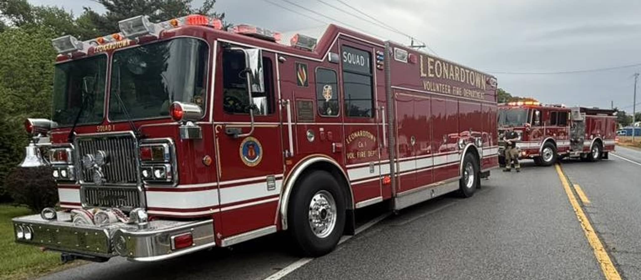 A red fire truck from Leonardtown Volunteer Fire Department parked on a road with additional emergency vehicles visible in the background under an overcast sky.