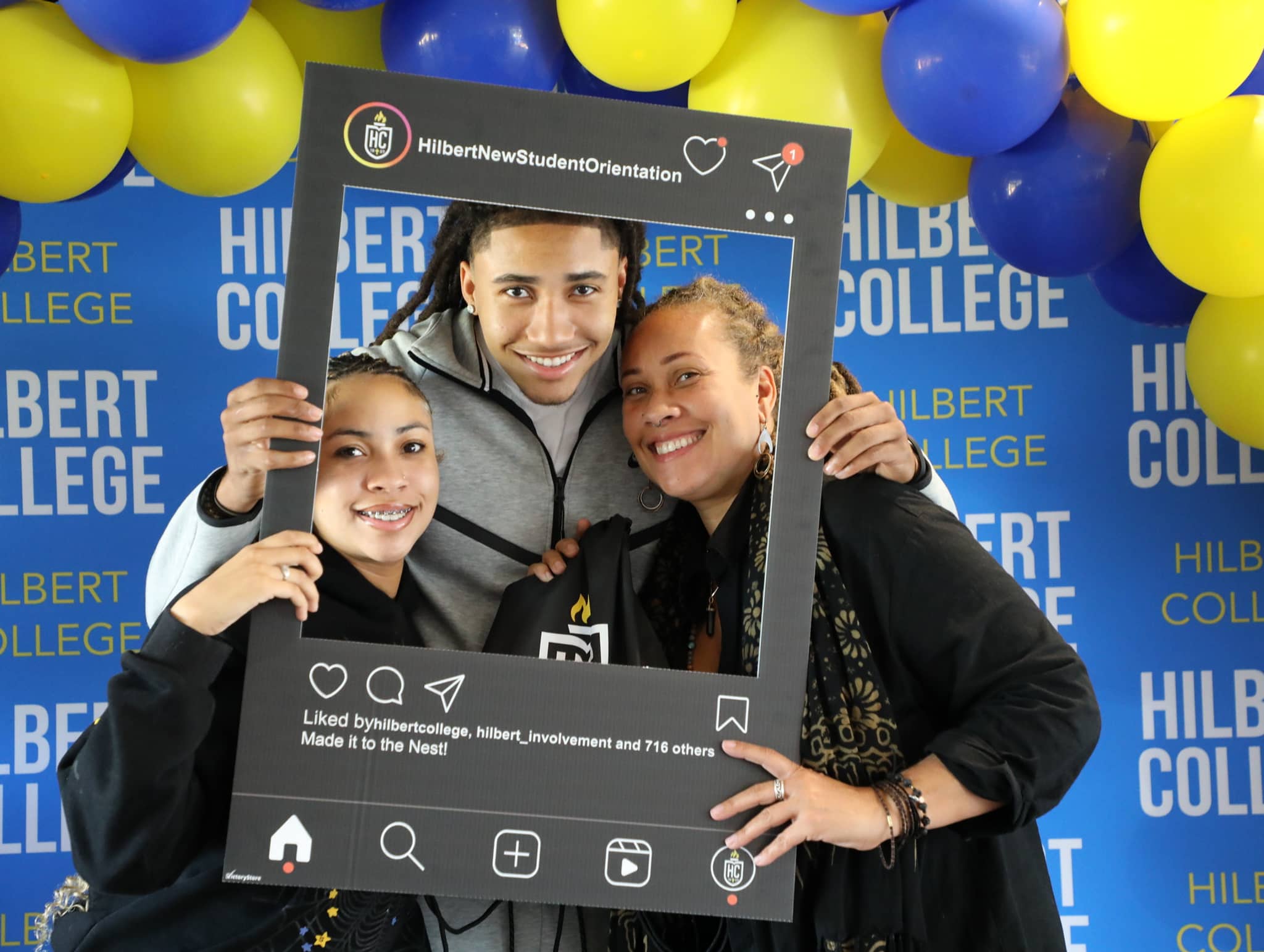 Four smiling students pose together holding a large Instagram-themed photo frame prop at Hilbert College's New Student Orientation event, surrounded by blue and yellow balloons and branded backdrop.