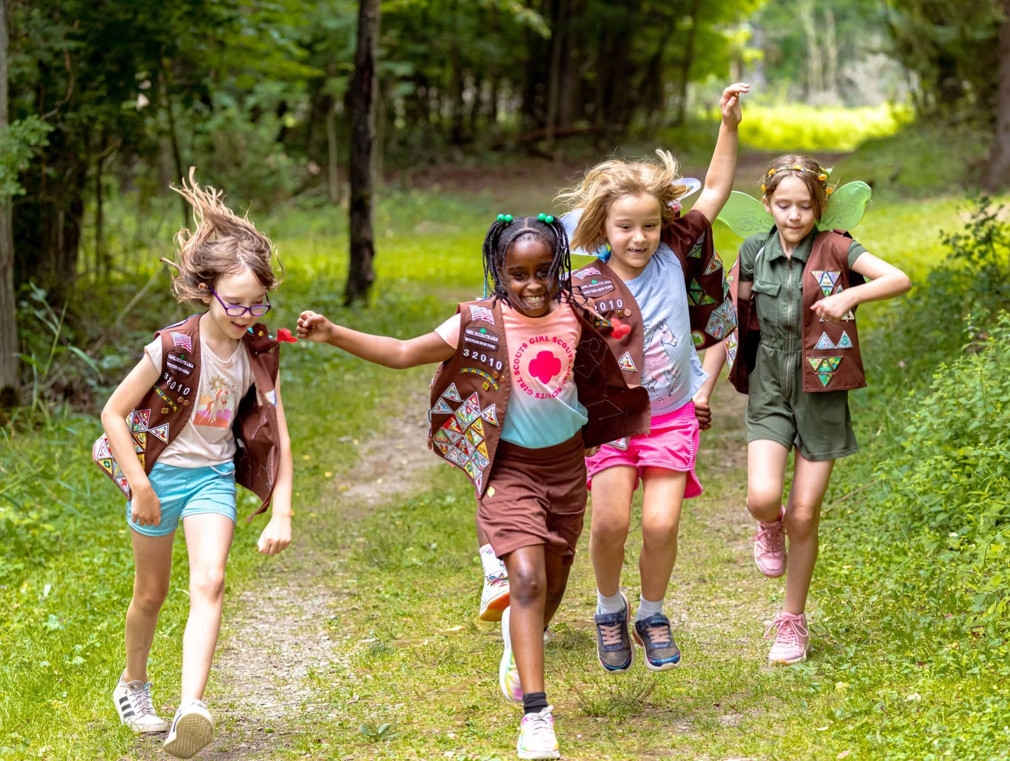 A group of Girl Scouts wearing brown vests with badges runs joyfully down a tree-lined forest path on a sunny day, displaying their enthusiasm and camaraderie.
