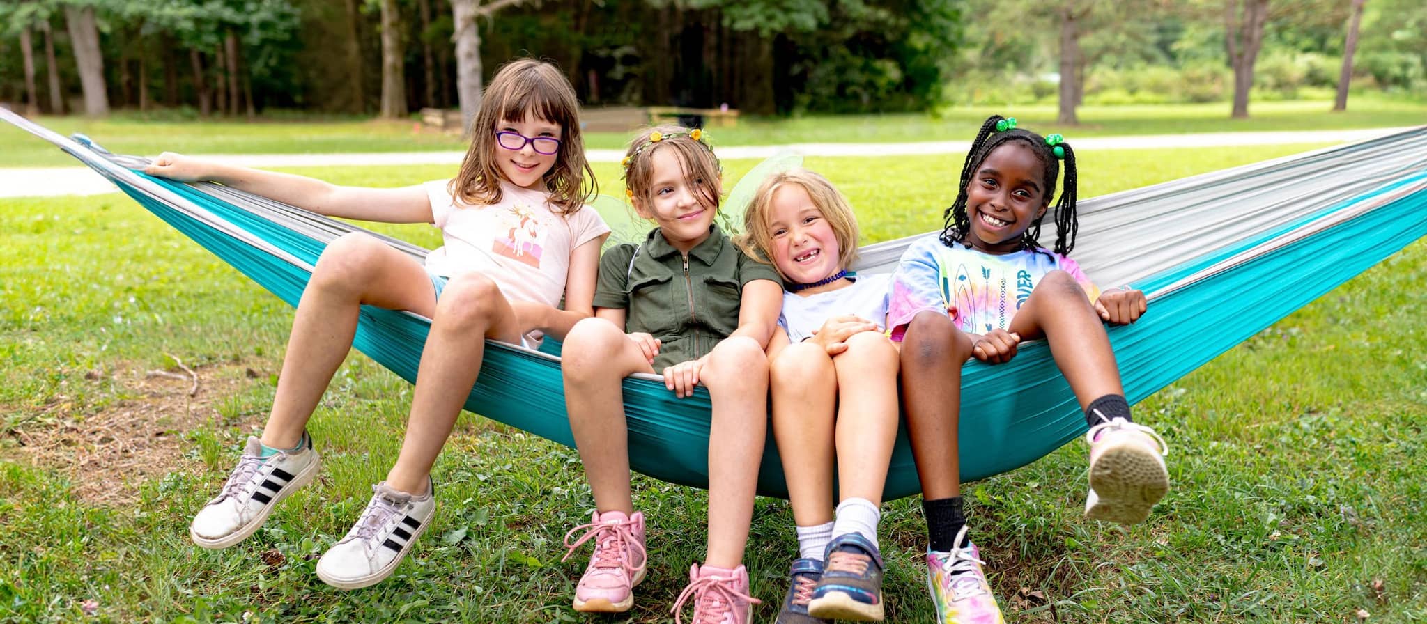 Four smiling children of different ethnicities sit together in a blue hammock in a grassy park with trees in the background, enjoying outdoor recreation.