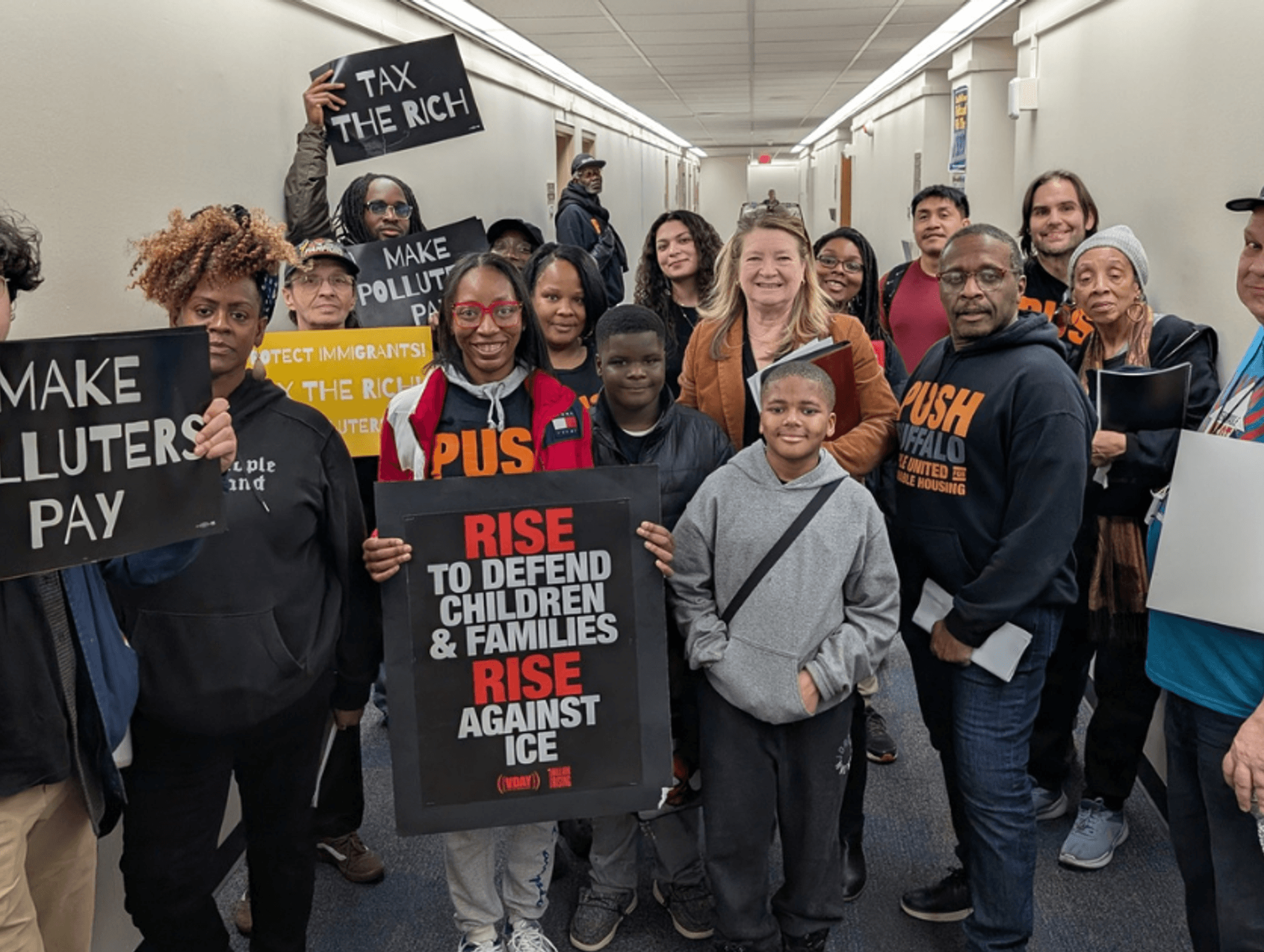 Group of diverse protesters gathered indoors holding signs advocating for taxing the wealthy, protecting immigrants, and defending families against ICE.