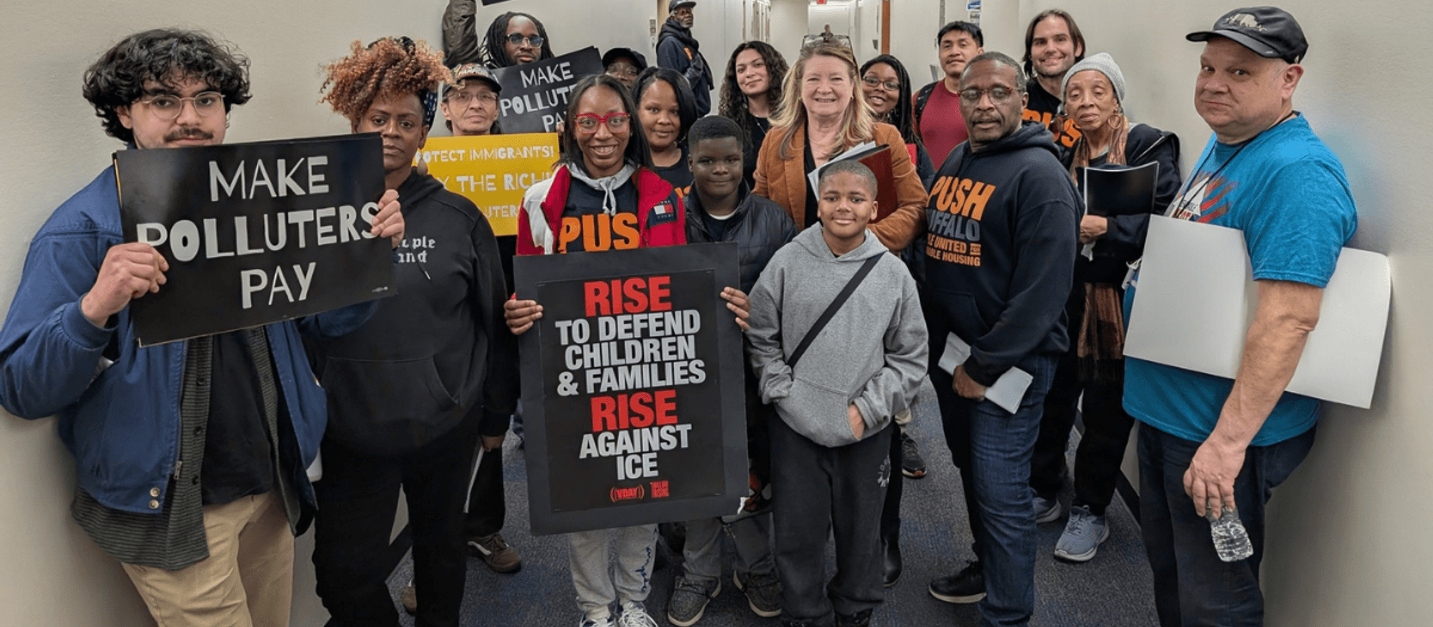 A diverse group of activists and protesters gather indoors holding signs with messages about taxing the rich, polluter accountability, immigrant protection, and defending families and children against ICE.