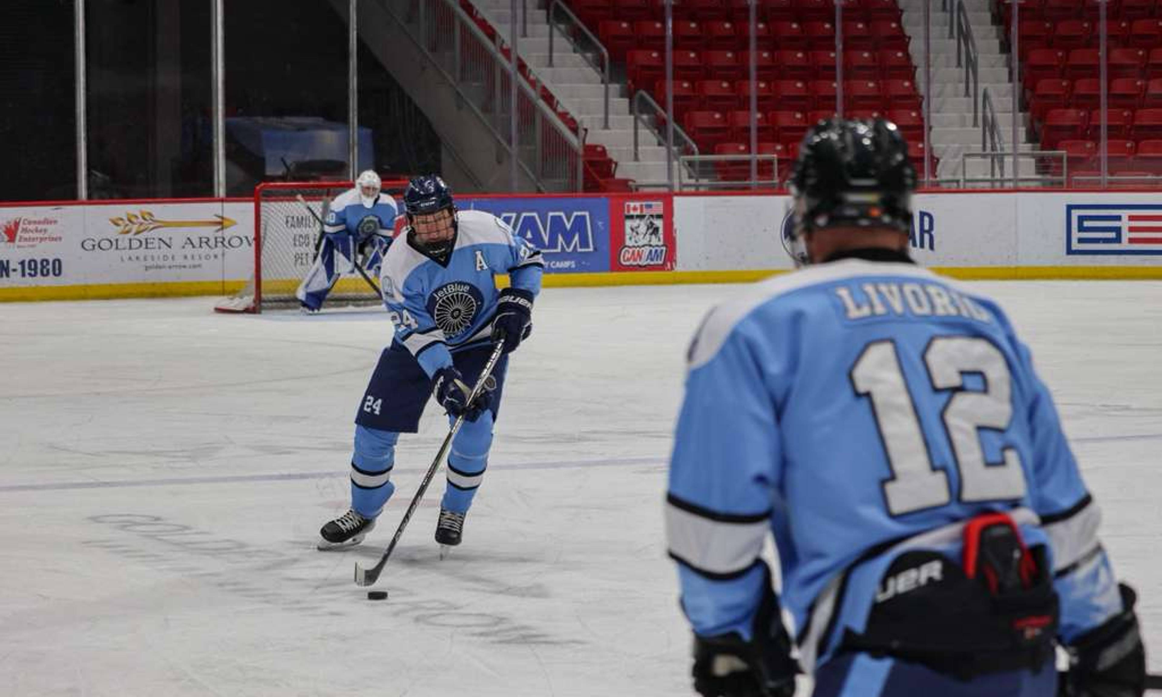 # Alt Text:

Hockey player in blue jersey number 24 controls the puck while teammate number 12 watches during a game in an arena with red seats and advertising boards.