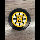 Boston Bruins hockey puck with team logo featuring yellow and black B with spoked wheel design, resting on gray wooden surface.