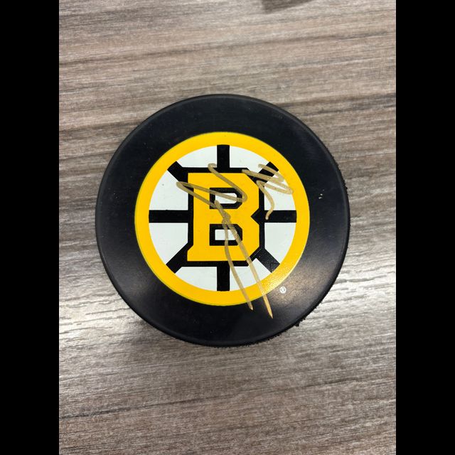 Boston Bruins hockey puck with team logo featuring yellow and black B with spoked wheel design, resting on gray wooden surface.