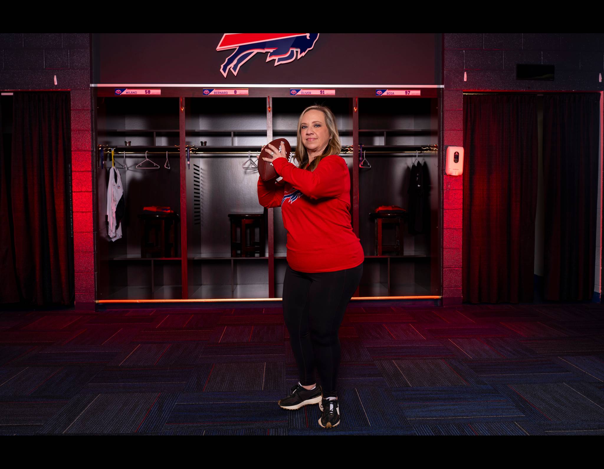 A woman in a red Bills jersey stands holding a football in the team's locker room with a Bills logo overhead.