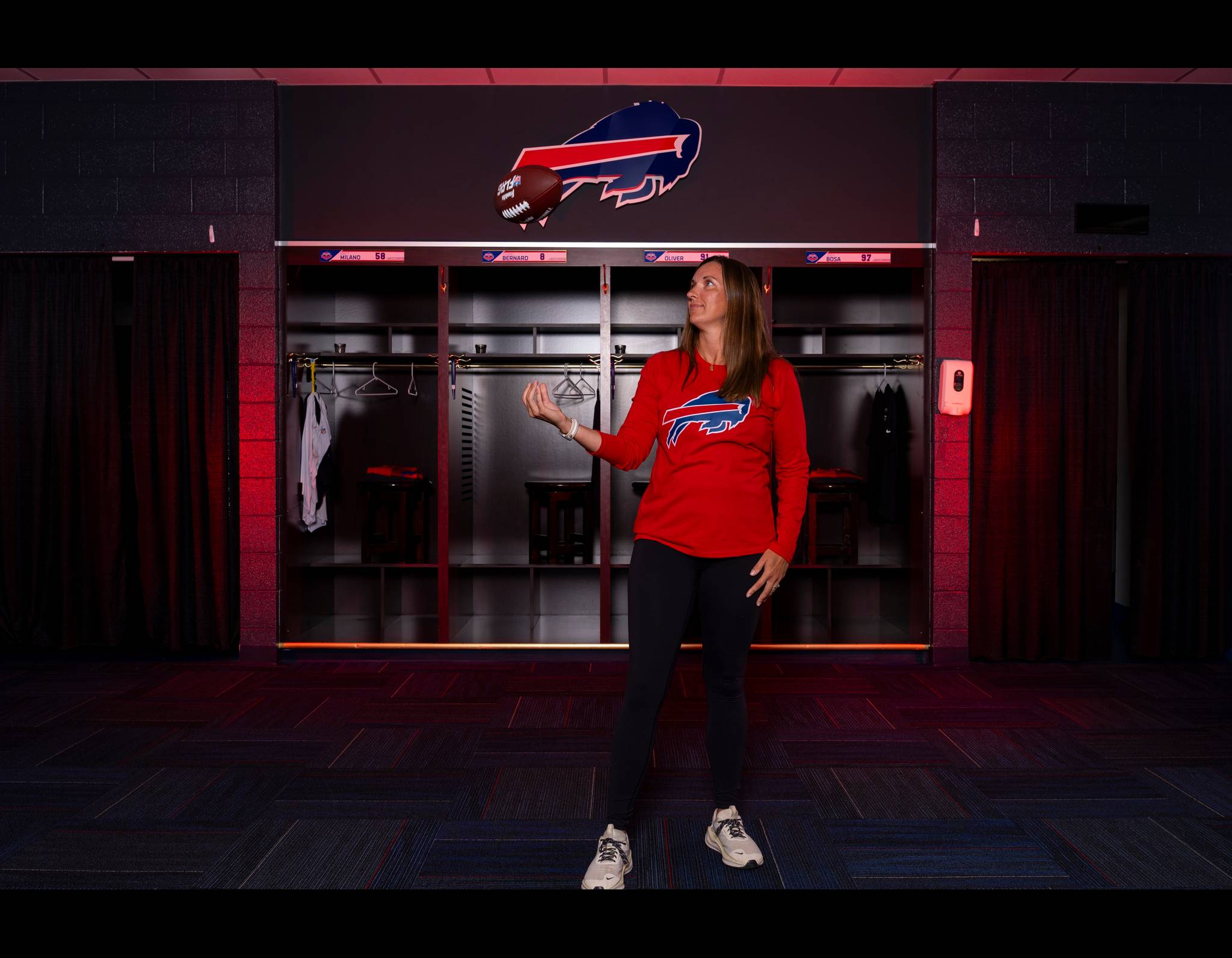 Woman in red Buffalo Bills shirt standing in team locker room with Bills logo in background, gesturing expressively.