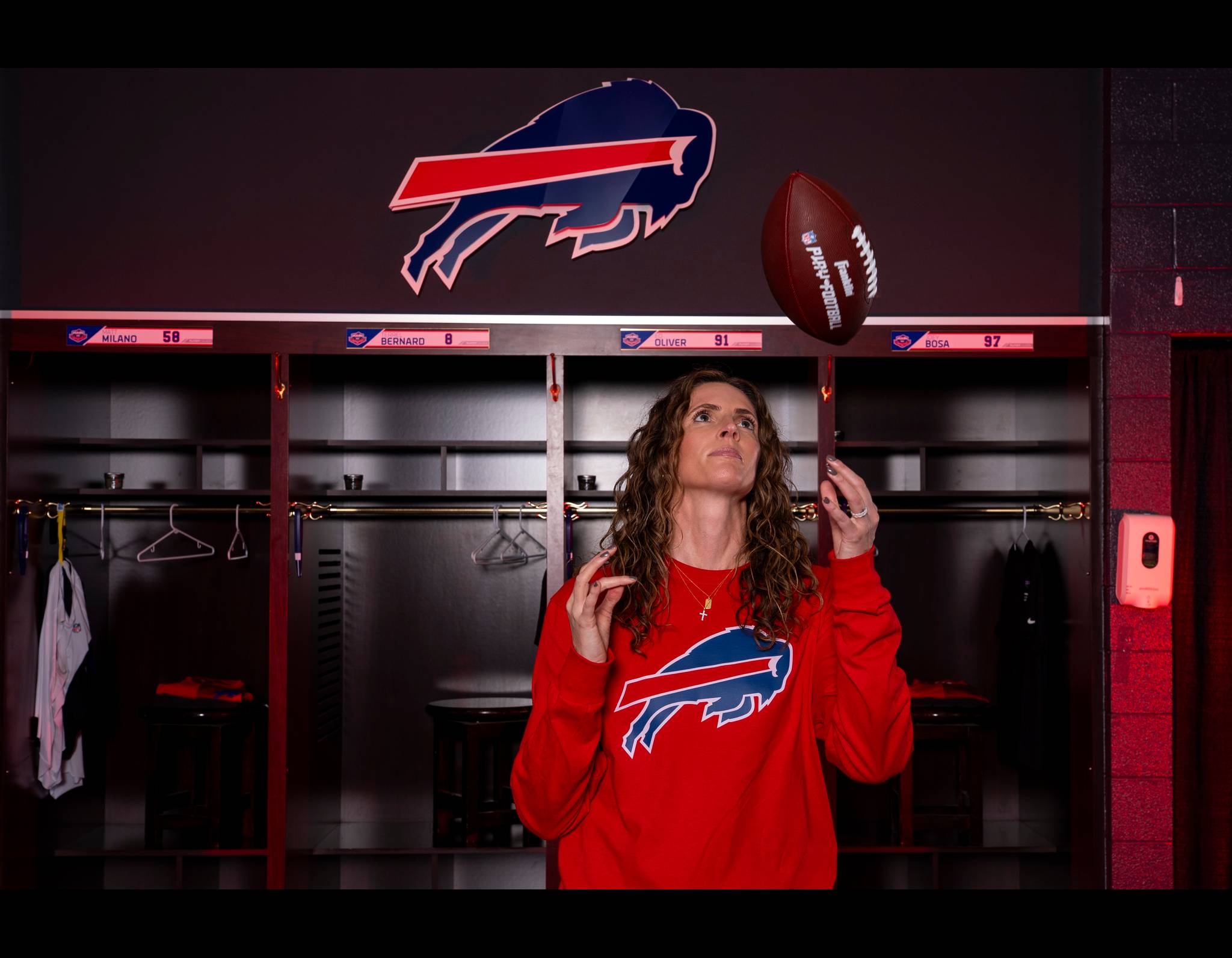 Woman in red Buffalo Bills sweatshirt looking up at football in Bills locker room with team logo in background