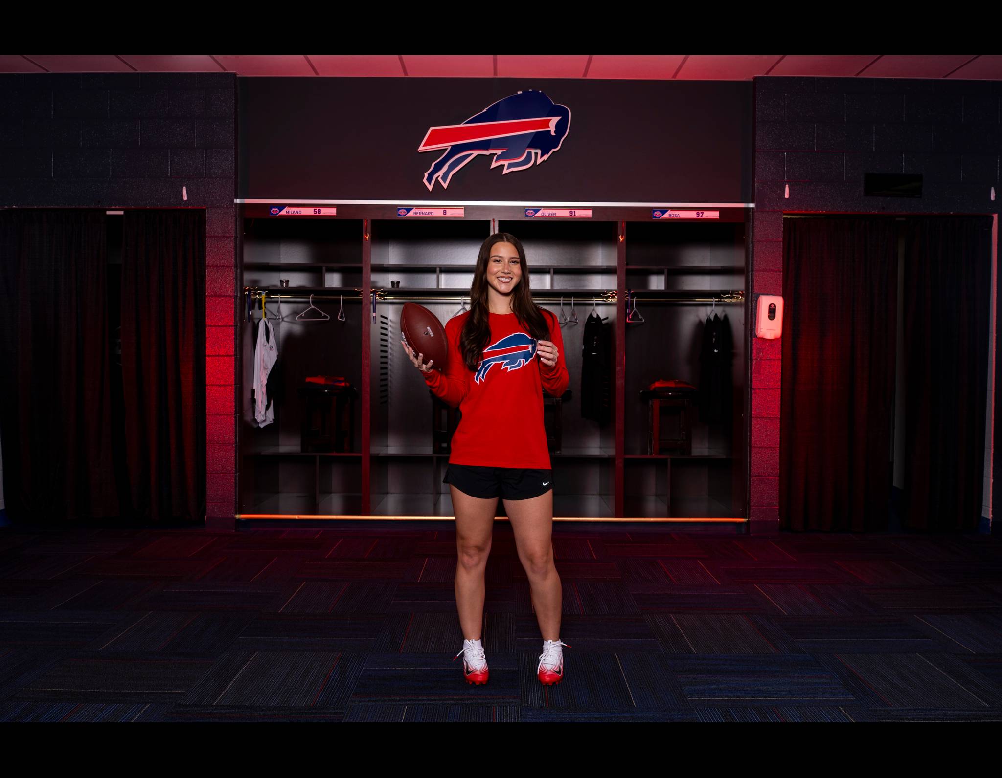 Woman in red Buffalo Bills shirt holding football, standing in team locker room with Bills logo behind her