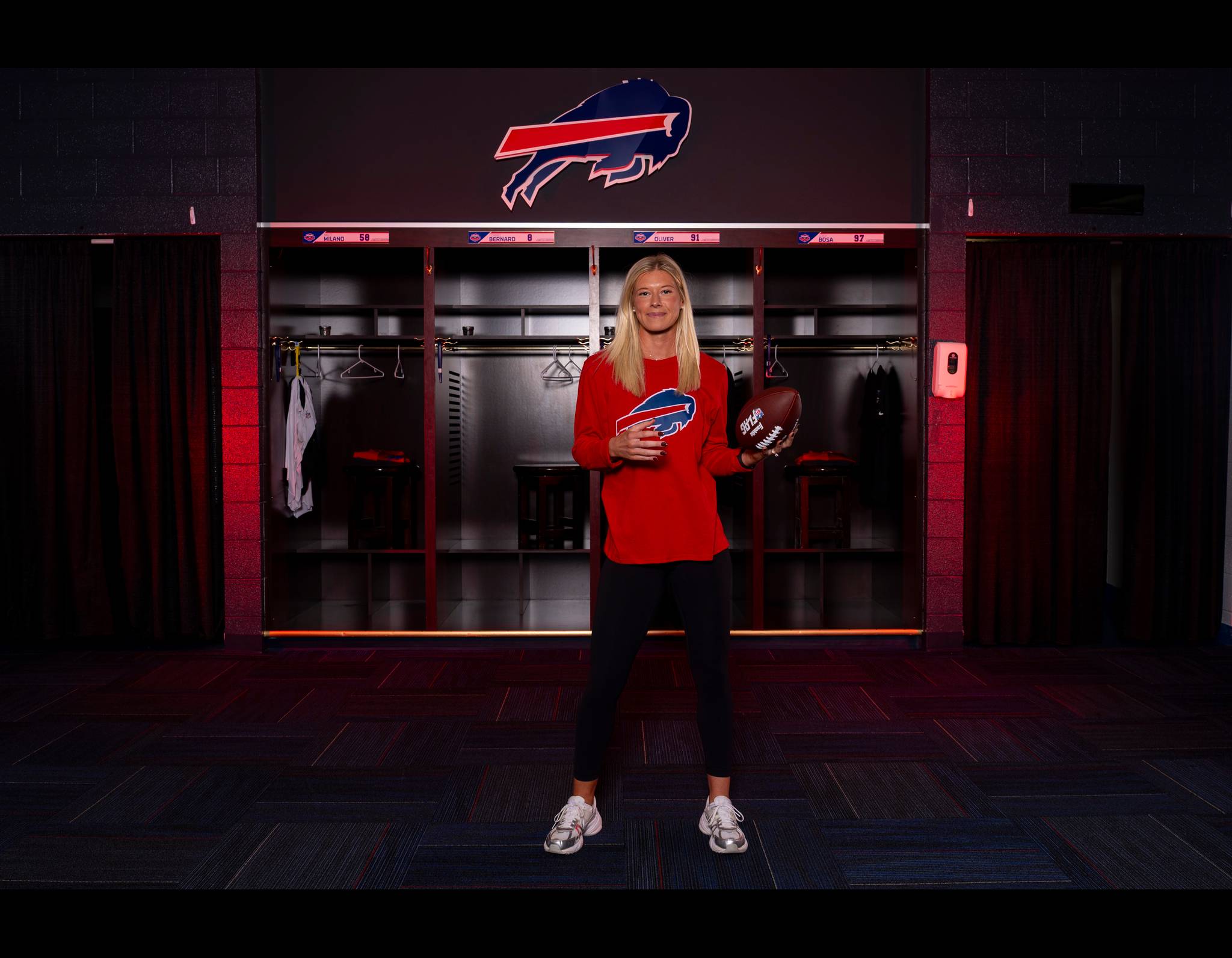 Blonde woman in red Buffalo Bills shirt holding football in team locker room with Bills logo above