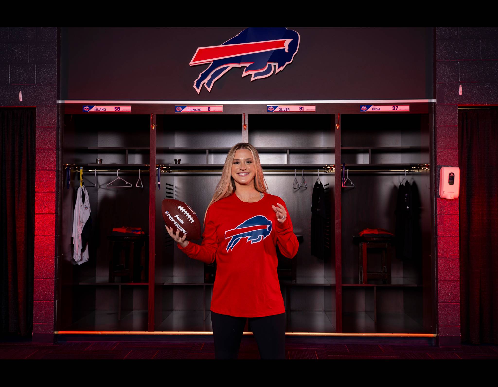 Smiling woman in red Bills shirt holding football in team locker room with player nameplates behind her