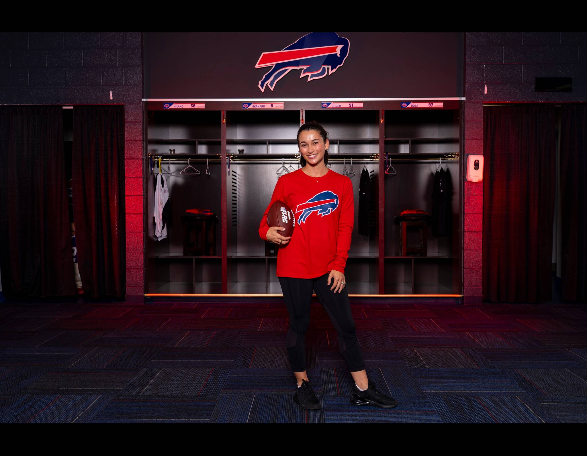 Smiling person in red Buffalo Bills shirt holding football in team locker room with team logo behind her