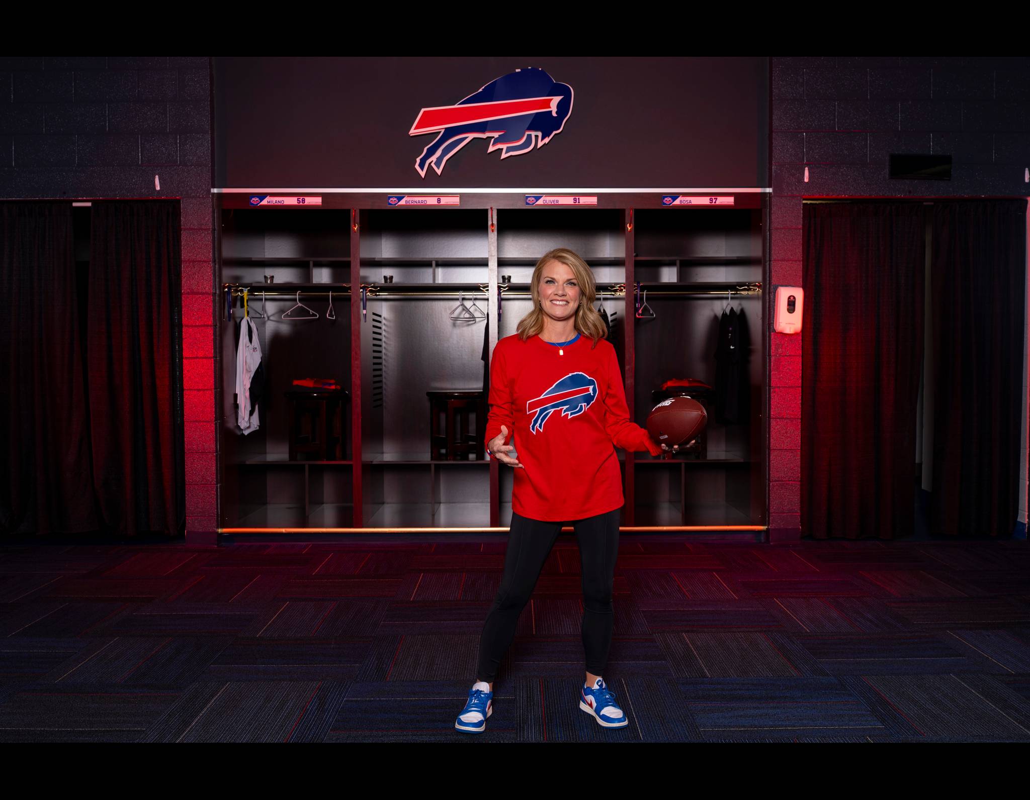 Woman in red Buffalo Bills sweater standing in team locker room, holding football, smiling brightly