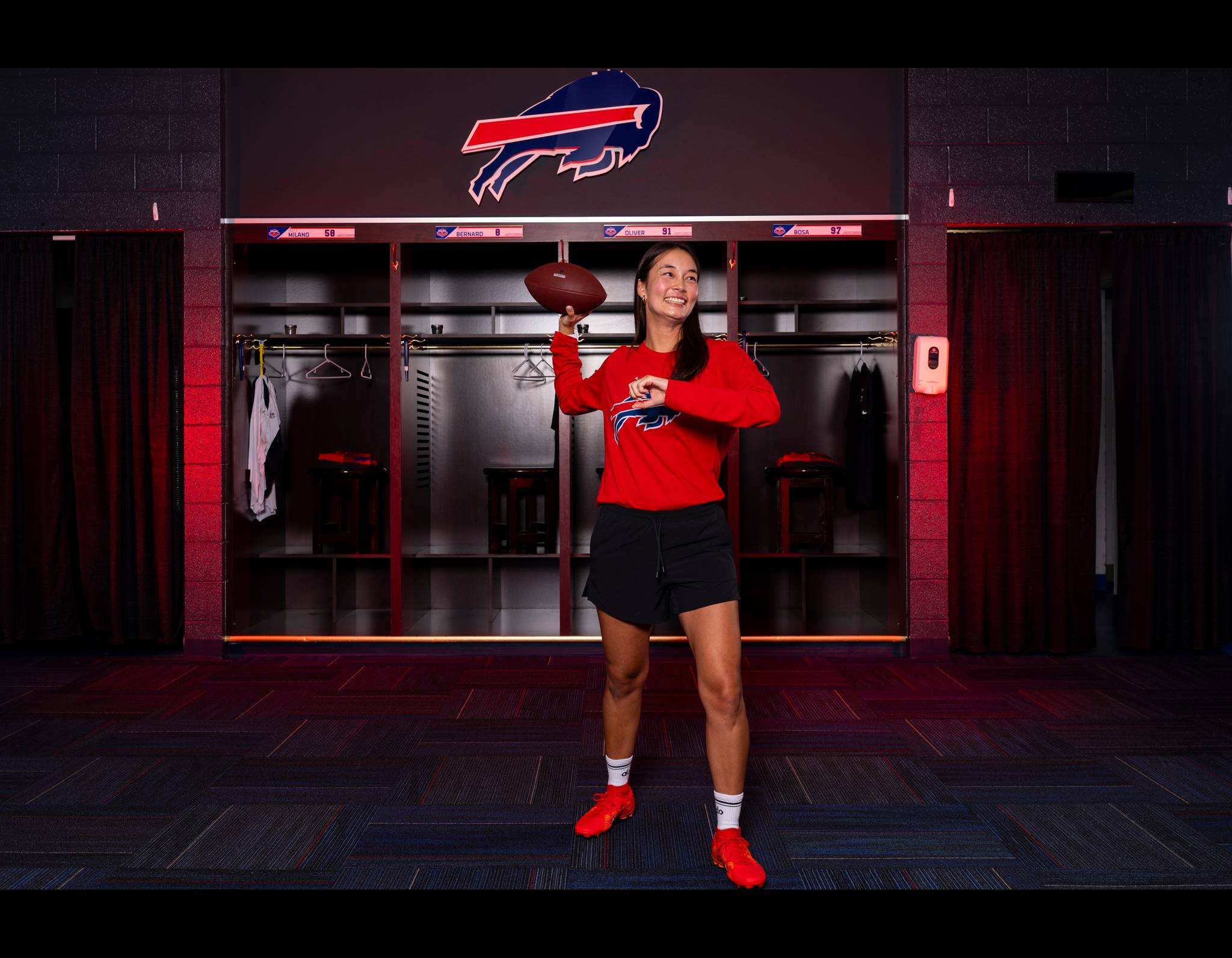 Smiling woman in red Bills sweatshirt holding football in team locker room with Buffalo Bills logo overhead