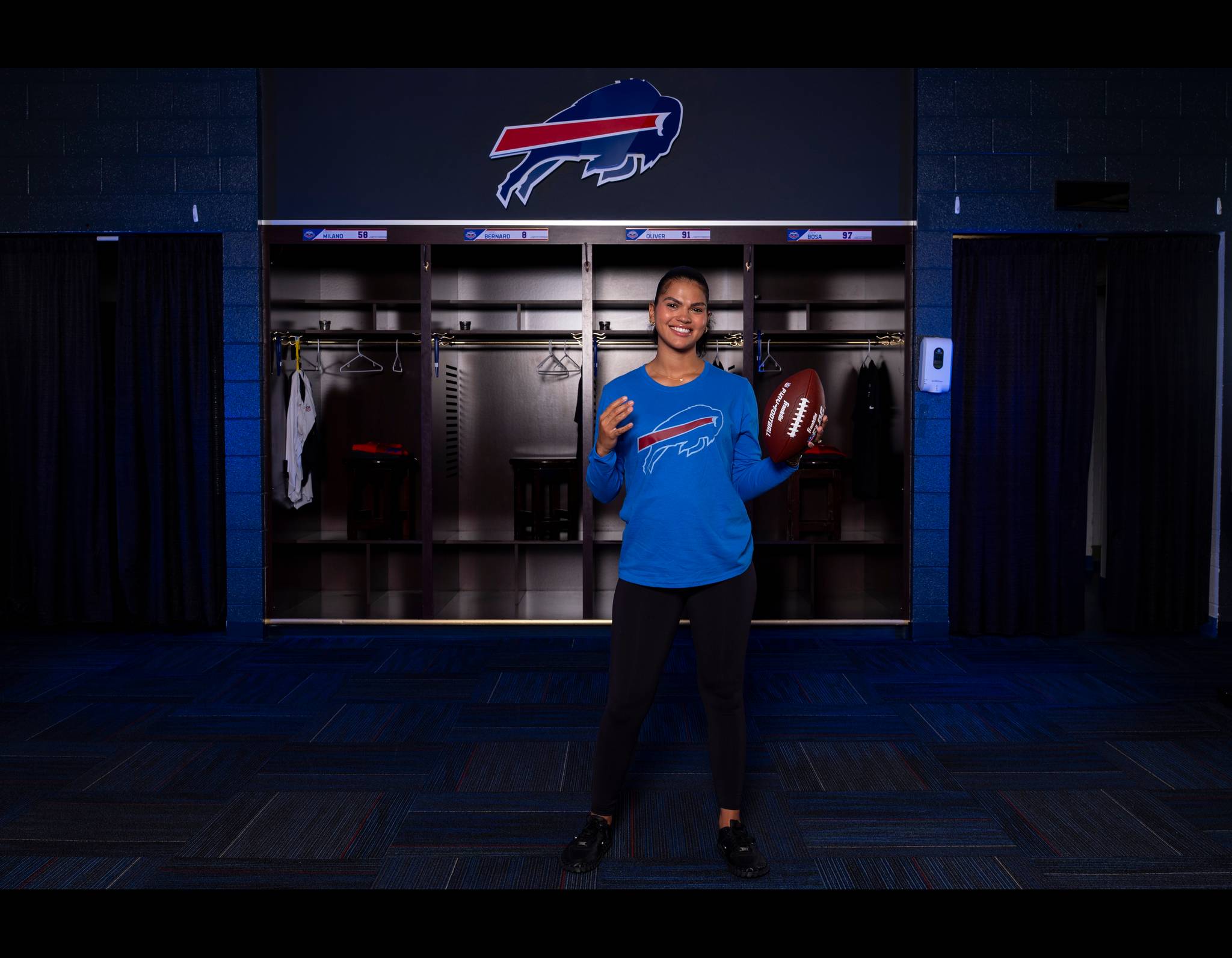 Smiling person in blue Bills shirt holding football, standing in front of team locker with Buffalo Bills logo overhead