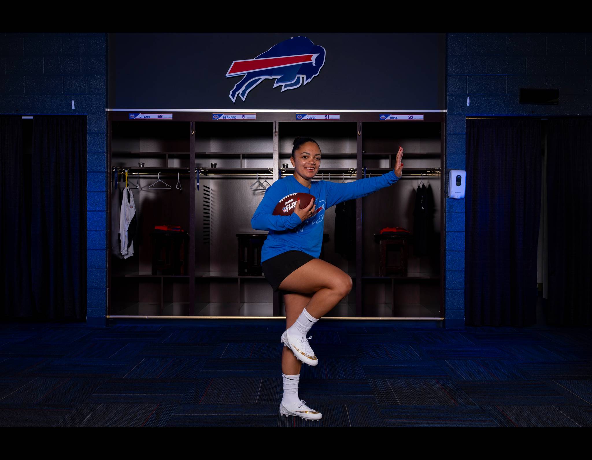 Smiling athlete in blue Bills shirt holds football, poses playfully in team locker room with team logo behind her