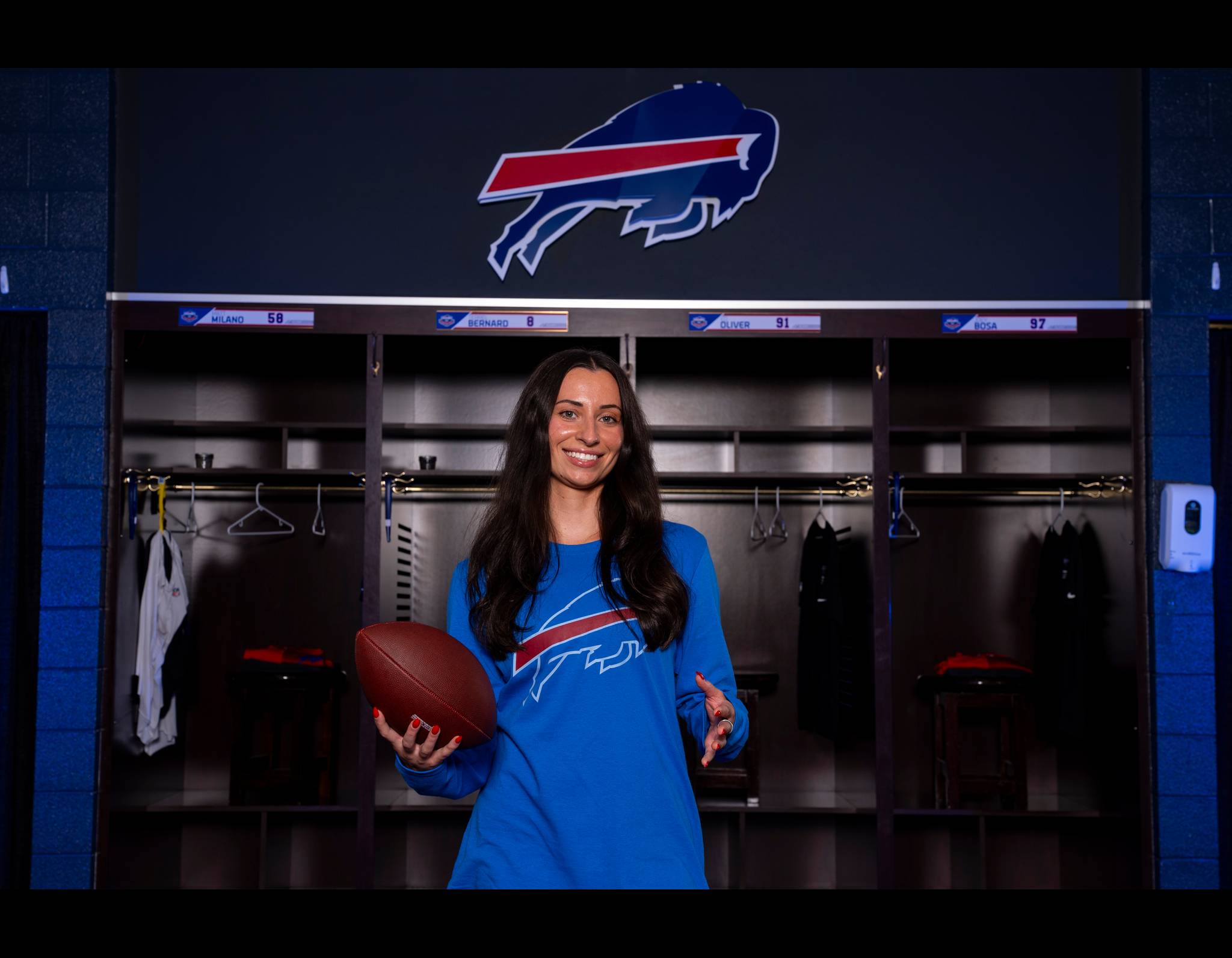 A woman in a blue Buffalo Bills shirt smiles in the team's locker room, holding a football in front of player lockers.