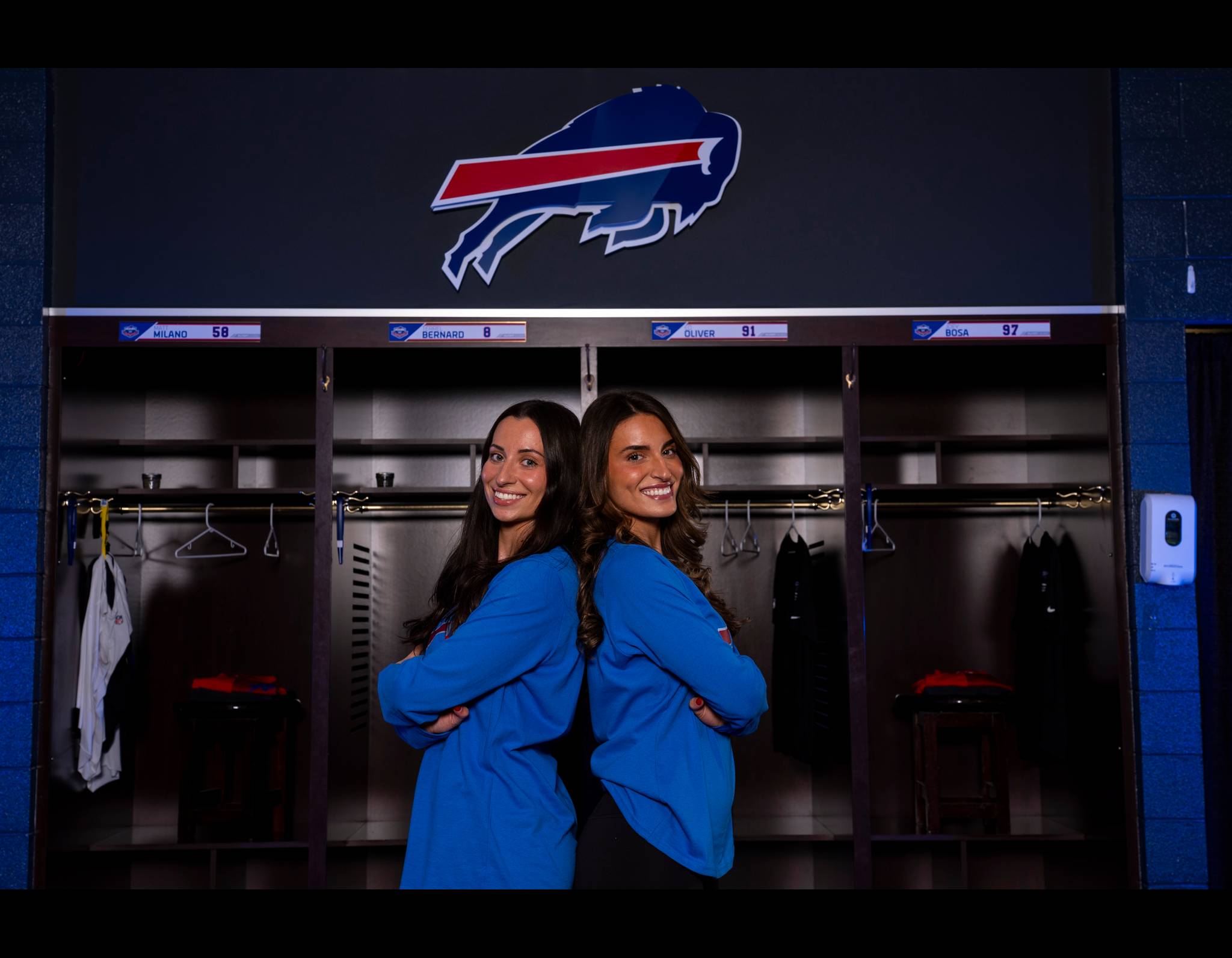 Two women in blue tops pose back-to-back in Buffalo Bills locker room with team logo and player lockers behind them.