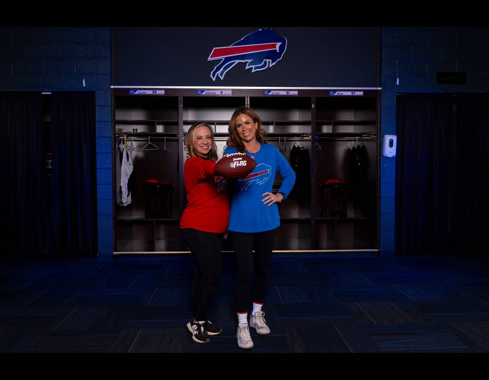 Two women posing with a football in front of Buffalo Bills lockers, wearing red and blue team-related tops.