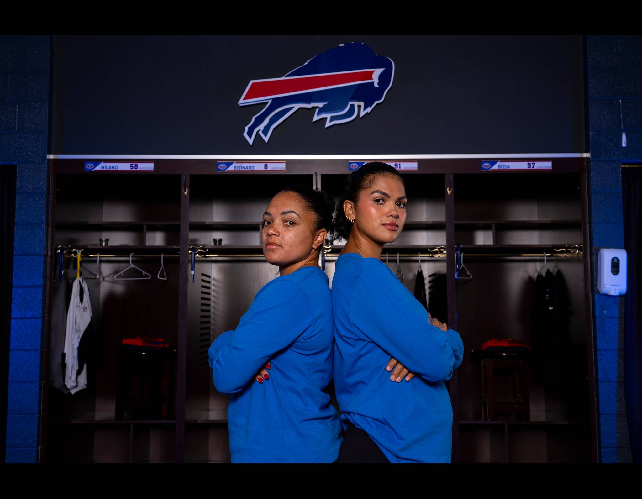 Two women in blue shirts stand back-to-back in Buffalo Bills locker room with team logo behind them