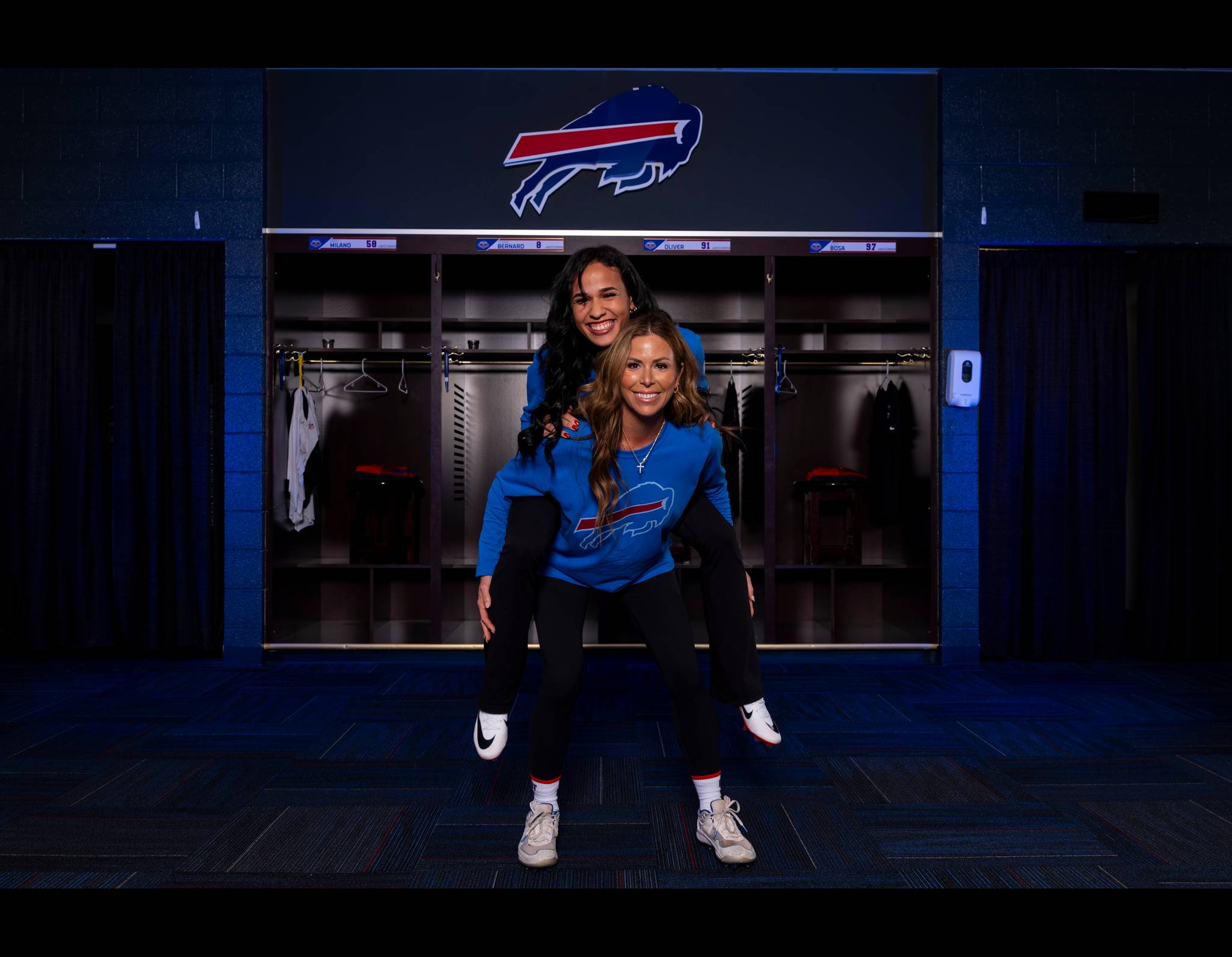 Two women in Buffalo Bills gear smiling in a team locker room, standing in front of open lockers and team logo