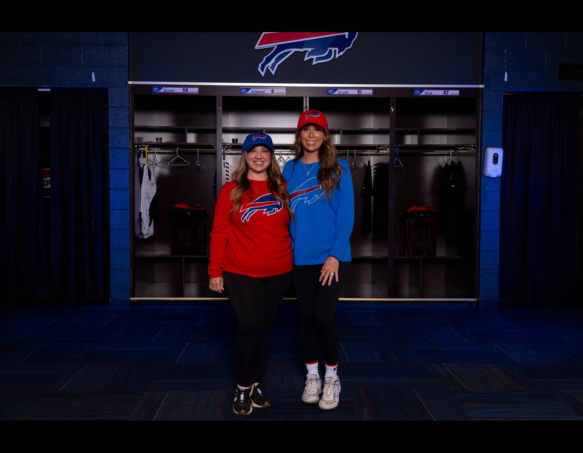 Two women in Buffalo Bills gear standing in team locker room, one in red shirt, one in blue, smiling together.