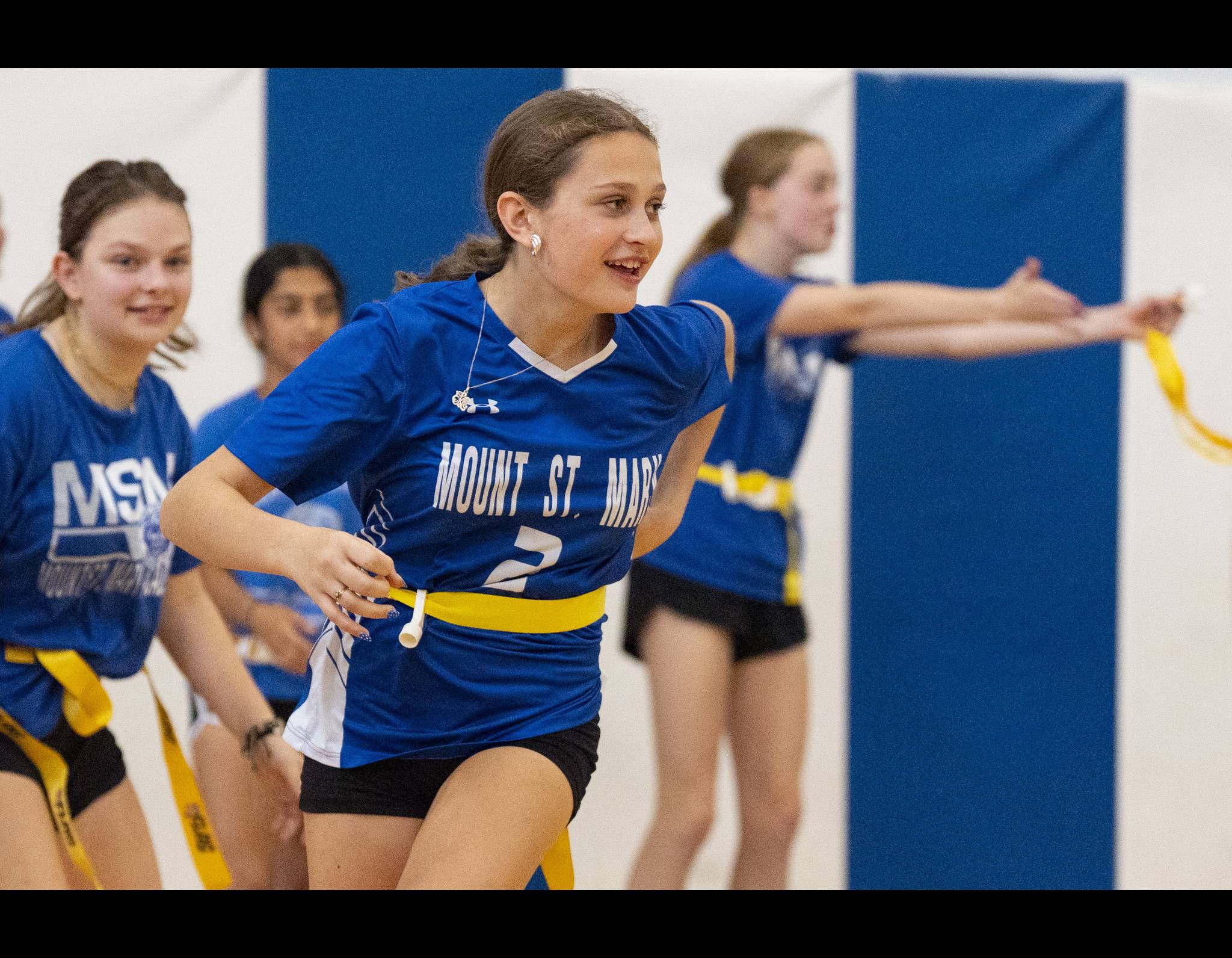 Blue-shirted student athletes playing flag football, smiling and running in a gymnasium with blue walls.