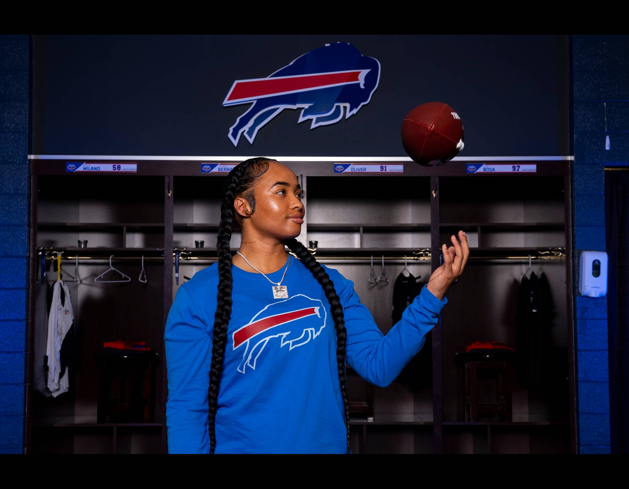 Buffalo Bills team member in blue jersey stands in locker room with football, wearing long braids and necklace.
