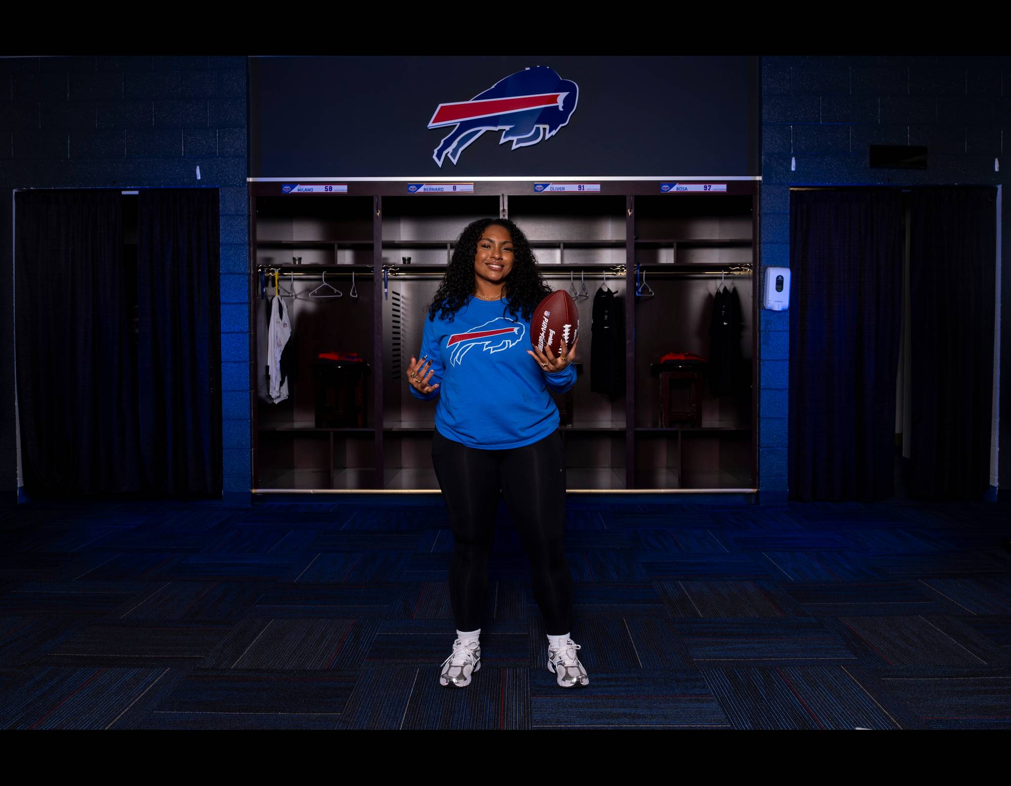 Person in blue Buffalo Bills shirt holding football in team locker room, standing in front of open lockers and team logo