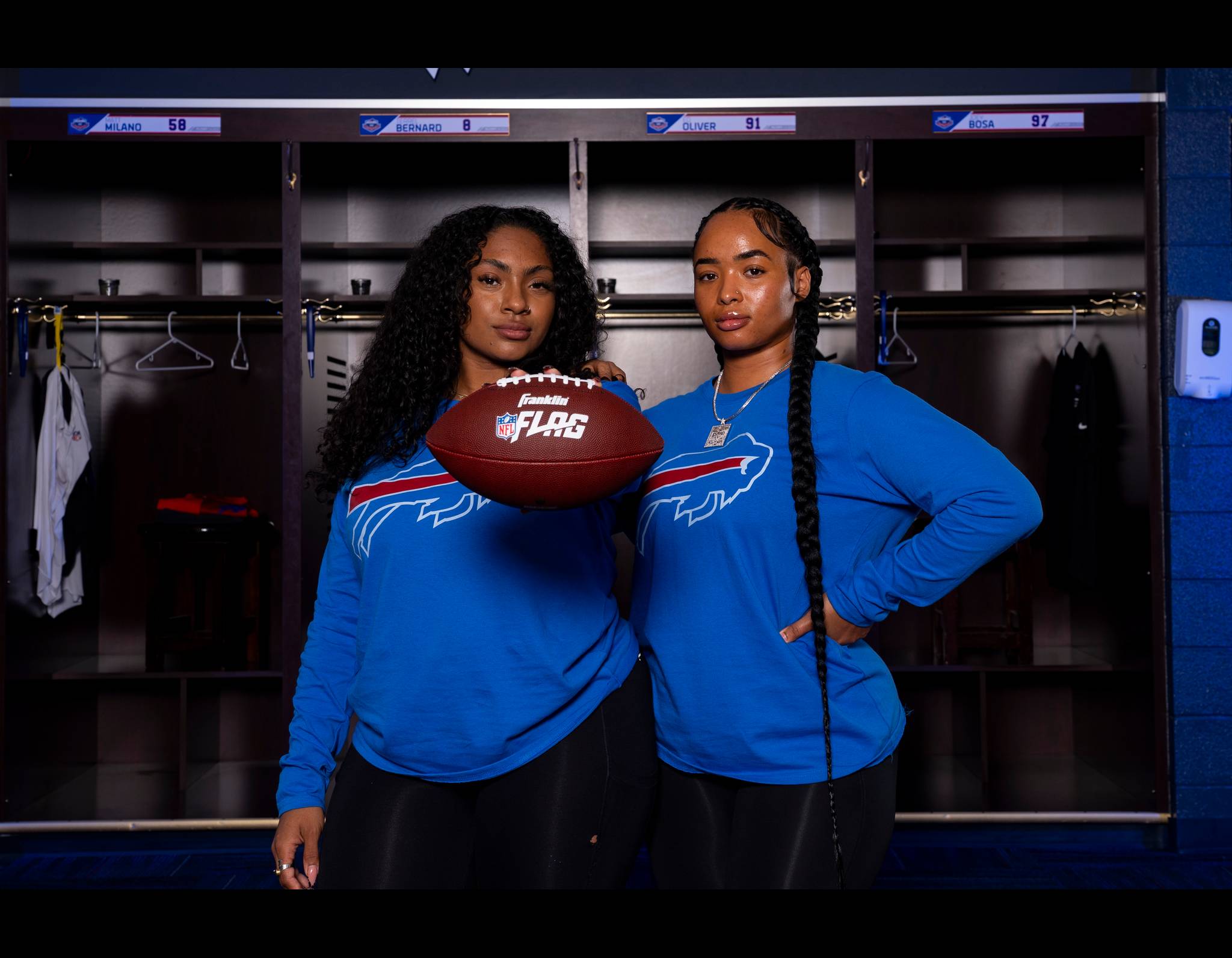 Two women in blue NFL Flag football shirts pose confidently in a locker room, holding a football between them.