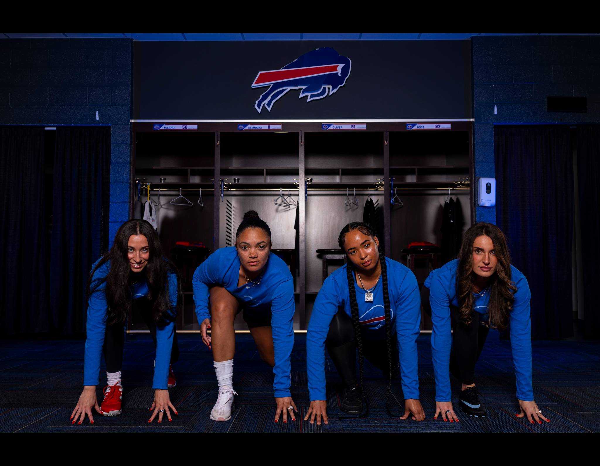 Four women in blue tops in athletic starting position in front of Buffalo Bills locker room, hands on ground, ready to sprint.
