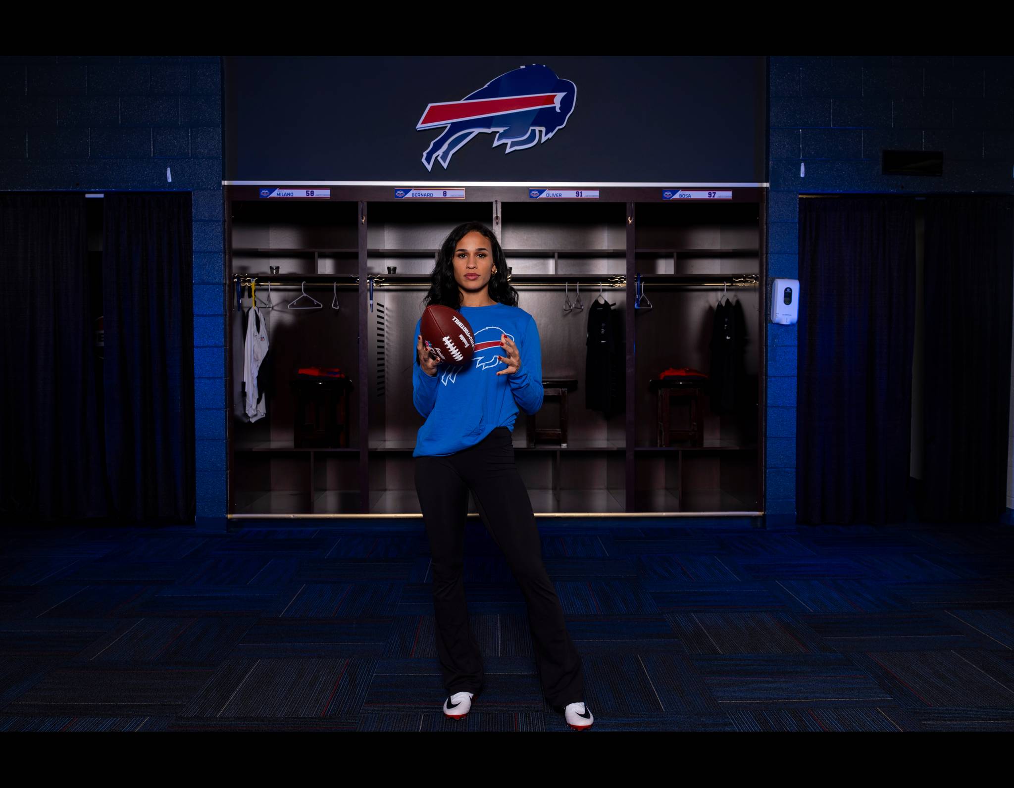 Woman in blue Bills sweatshirt holding football in team locker room with Buffalo Bills logo in background